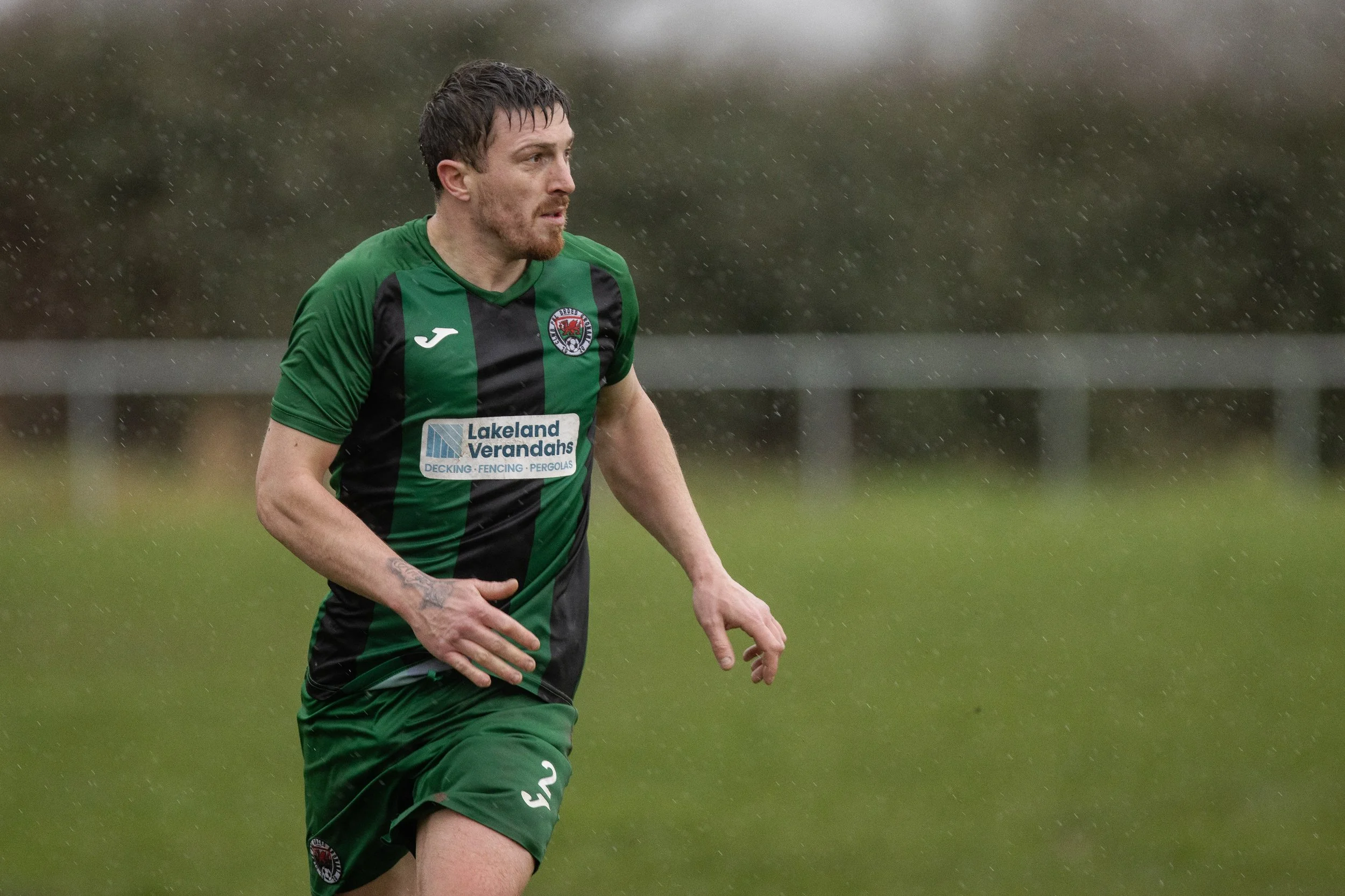 A male soccer player in a green and black uniform running in the rain on a grassy field.