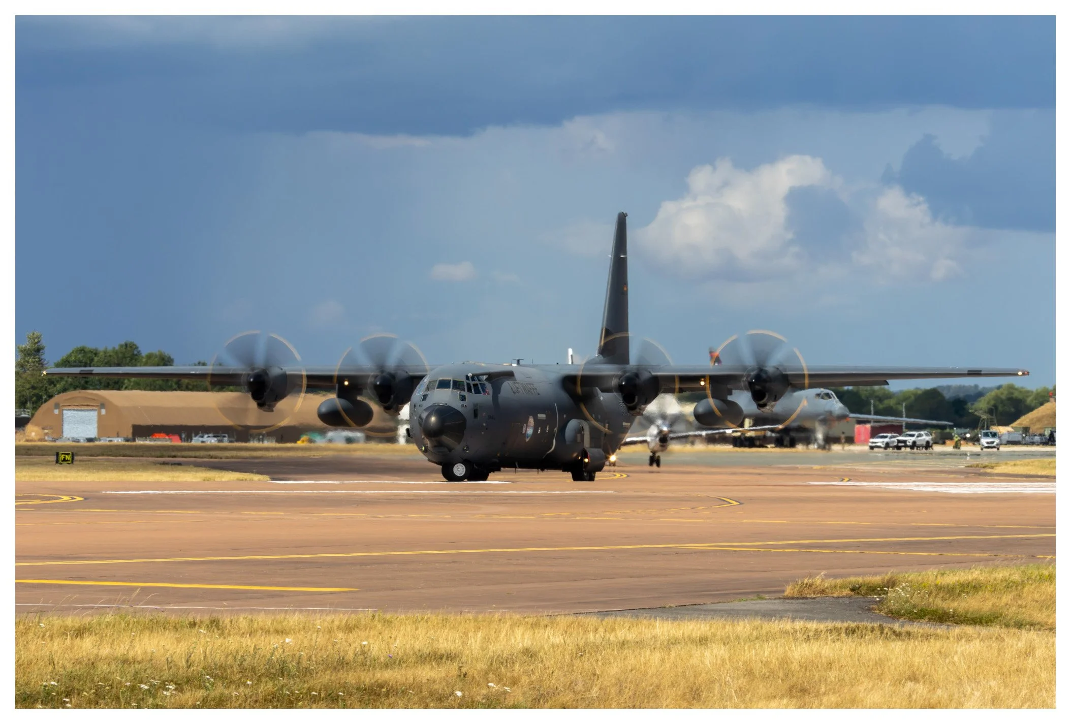 A military cargo airplane taxiing on the runway at an airport during daytime with other aircraft, vehicles, and airport structures in the background.