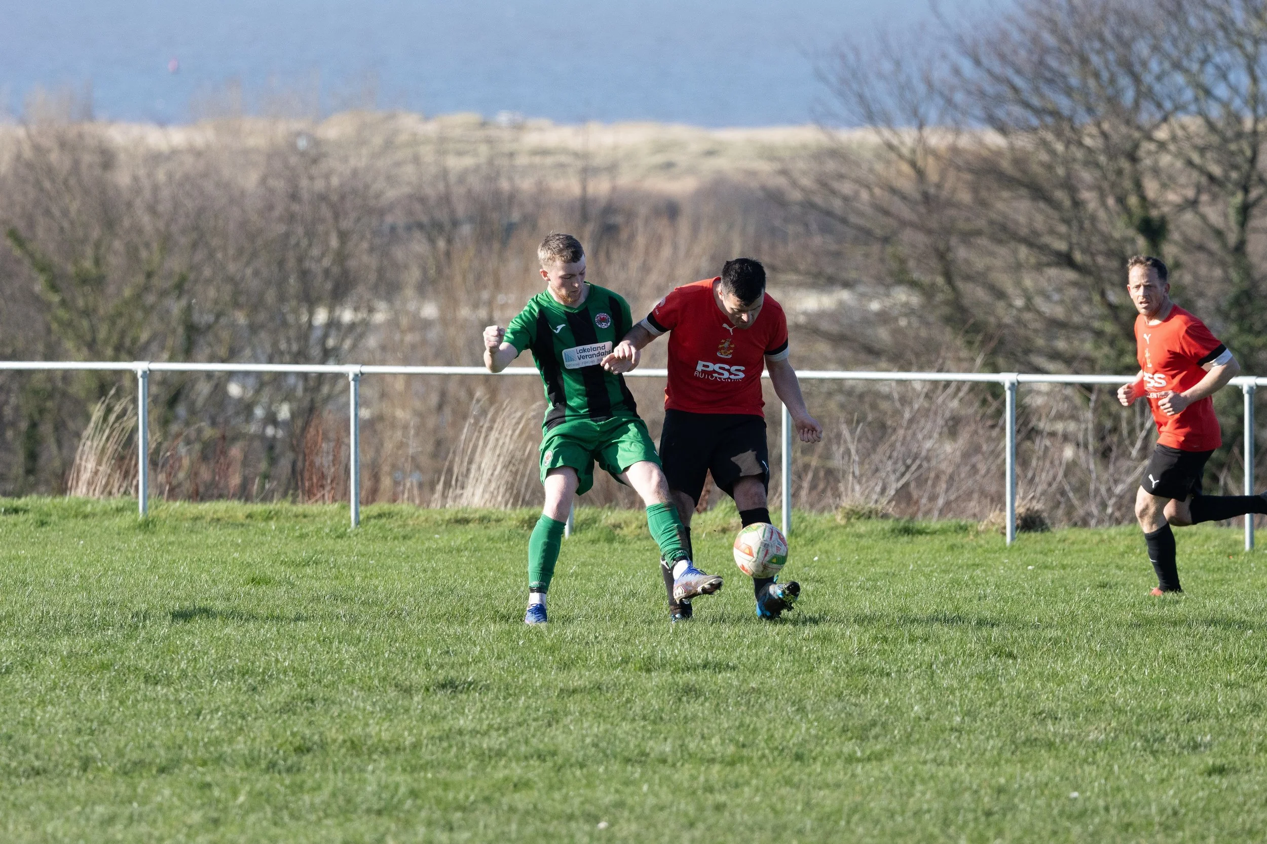 Soccer players in green and red uniforms compete for the ball on a grassy field with trees and water in the background.
