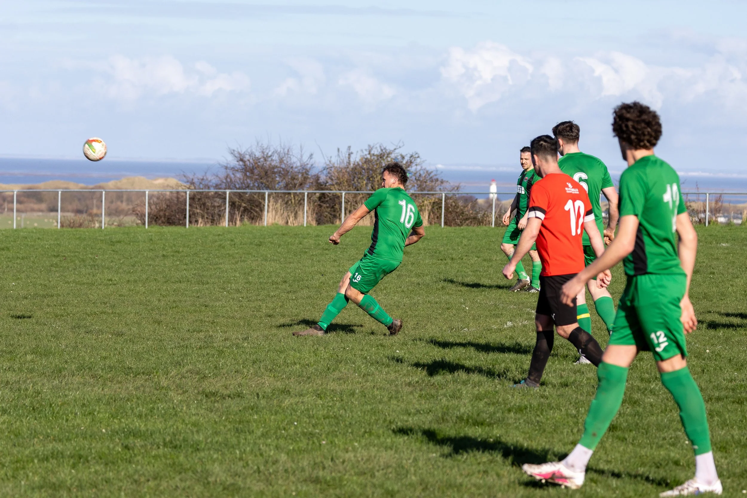 Soccer players wearing green and black uniforms playing on a grassy field, with one player in mid-kick and a soccer ball in the air.