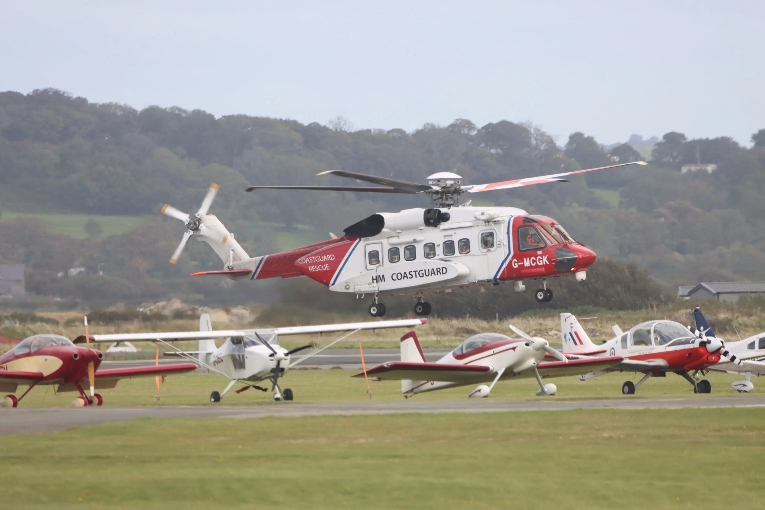 Colorful helicopter flying low over a grassy field with small airplanes parked on the ground and a hilly landscape in the background.