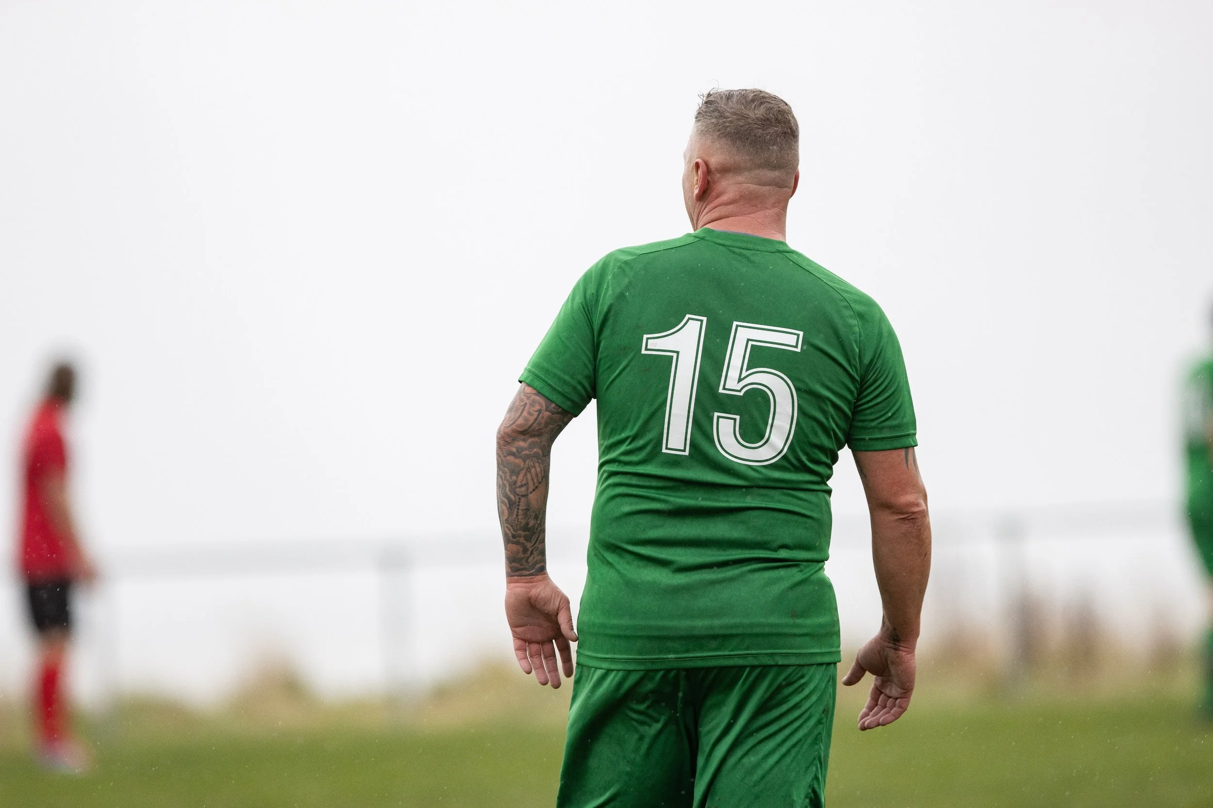 A man wearing a green sports jersey with the number 15 on the back is standing on a field, with a blurred person in red and black in the background.