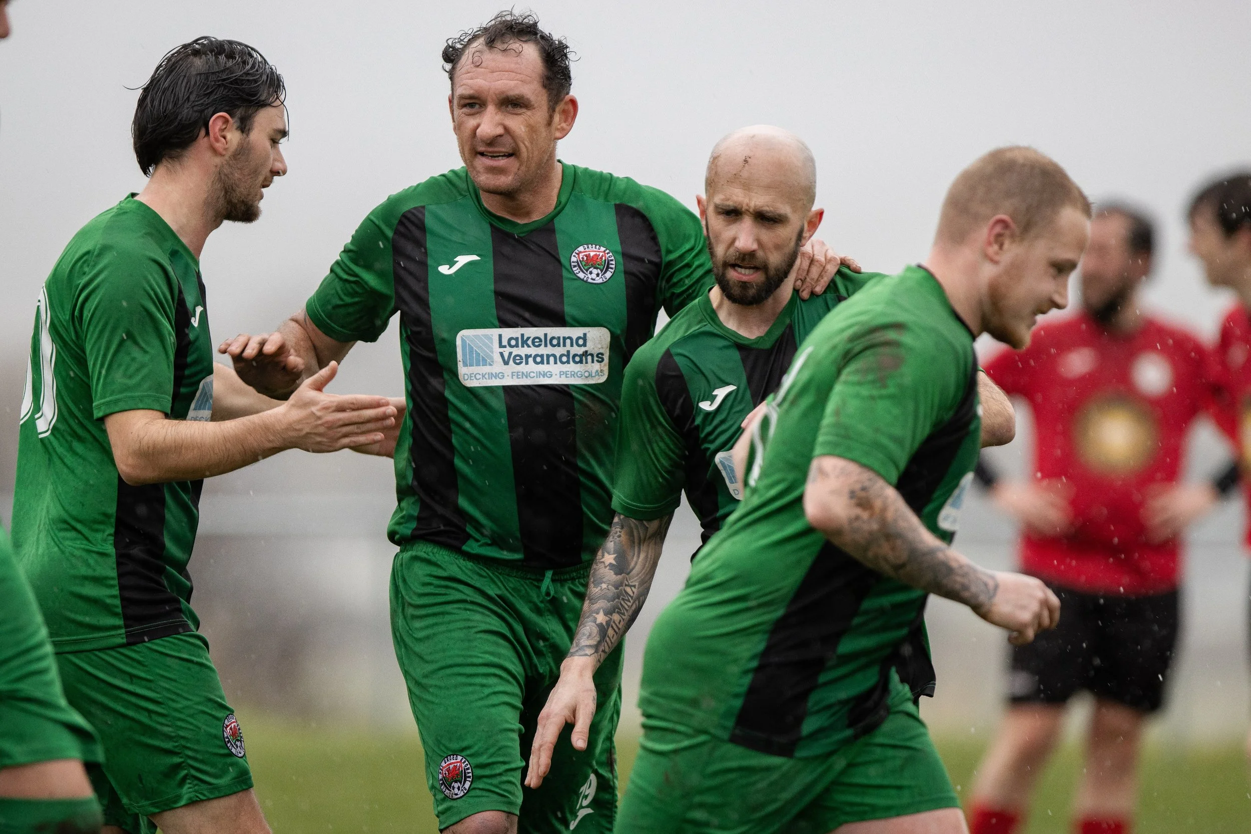 Soccer players in green and black uniforms celebrating on the field in rainy weather, with a blurred background of other players in red uniforms.