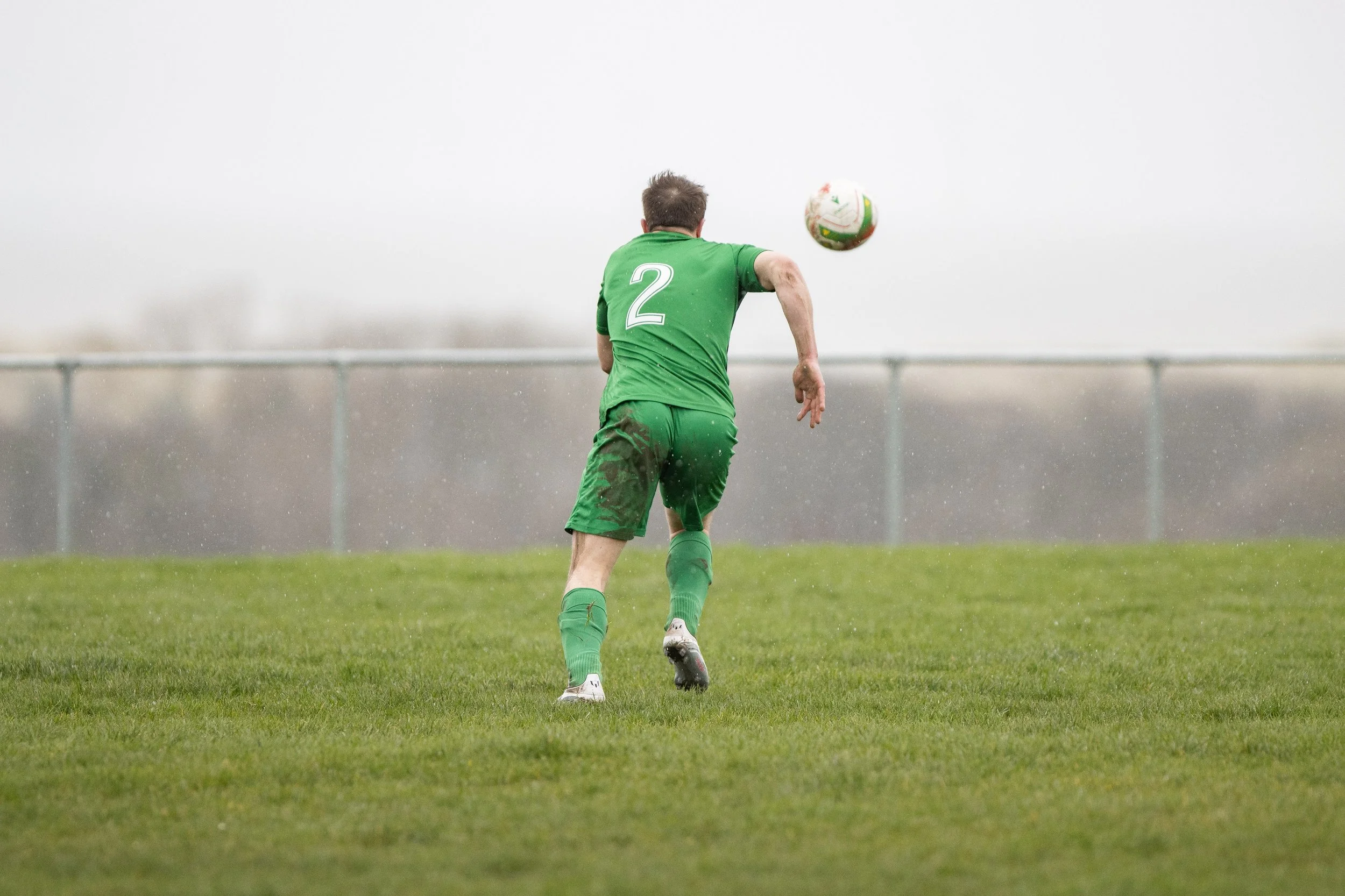 A soccer player in a green uniform with the number 2 on his back, running on a wet field while chasing a soccer ball.