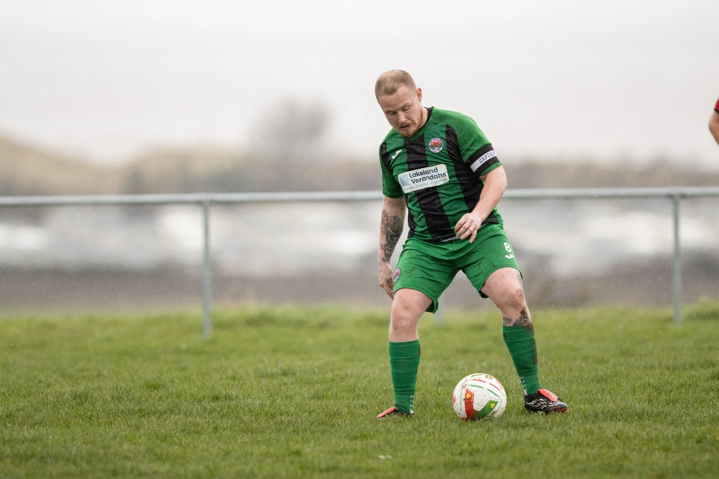 A soccer player in green and black uniform preparing to kick a soccer ball on a grassy field, with a blurred background and rain.