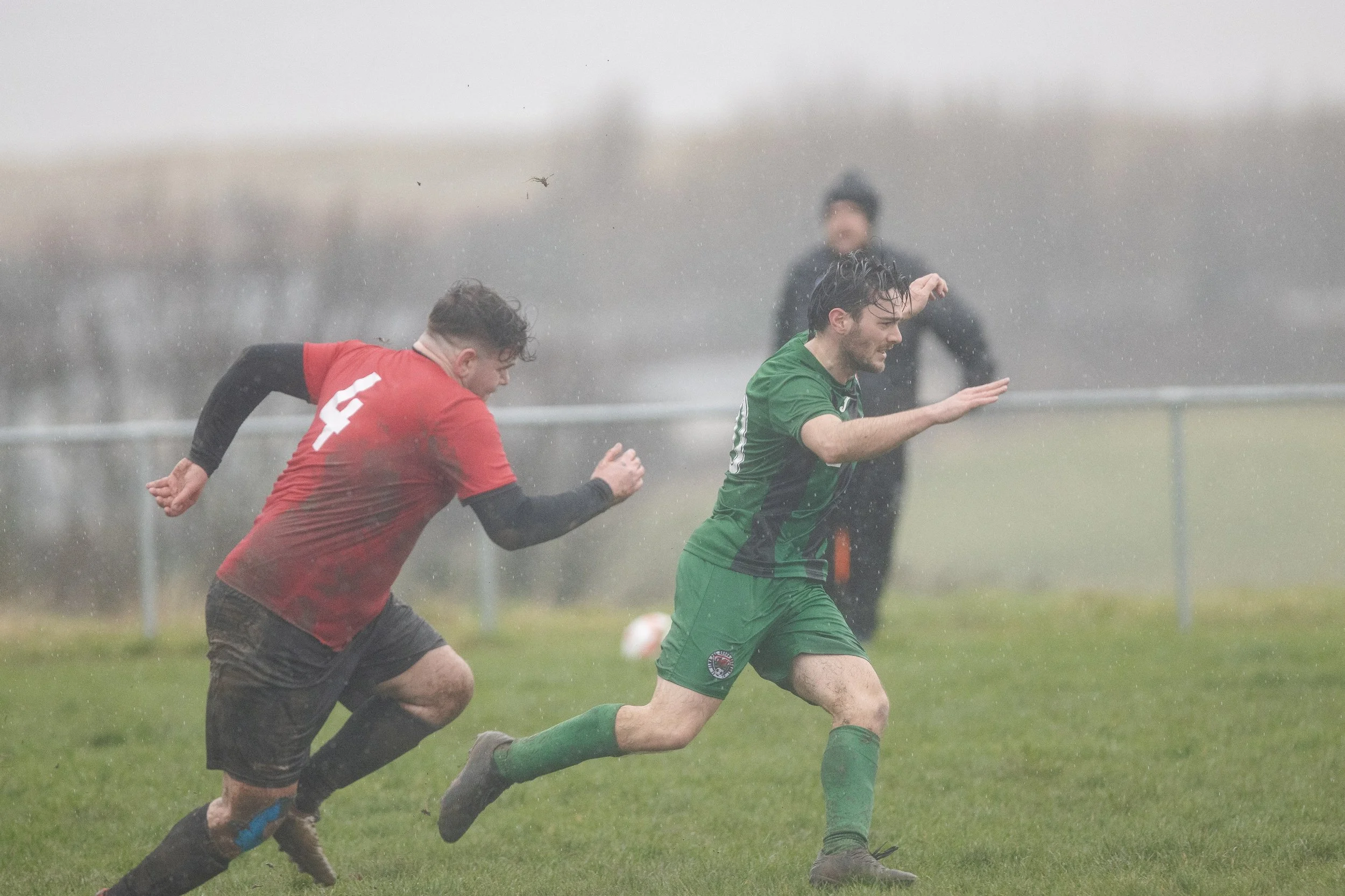 Two soccer players running in rain on a grassy field, one in a red shirt with the number 4 and the other in a green uniform, with a referee in black in the background.