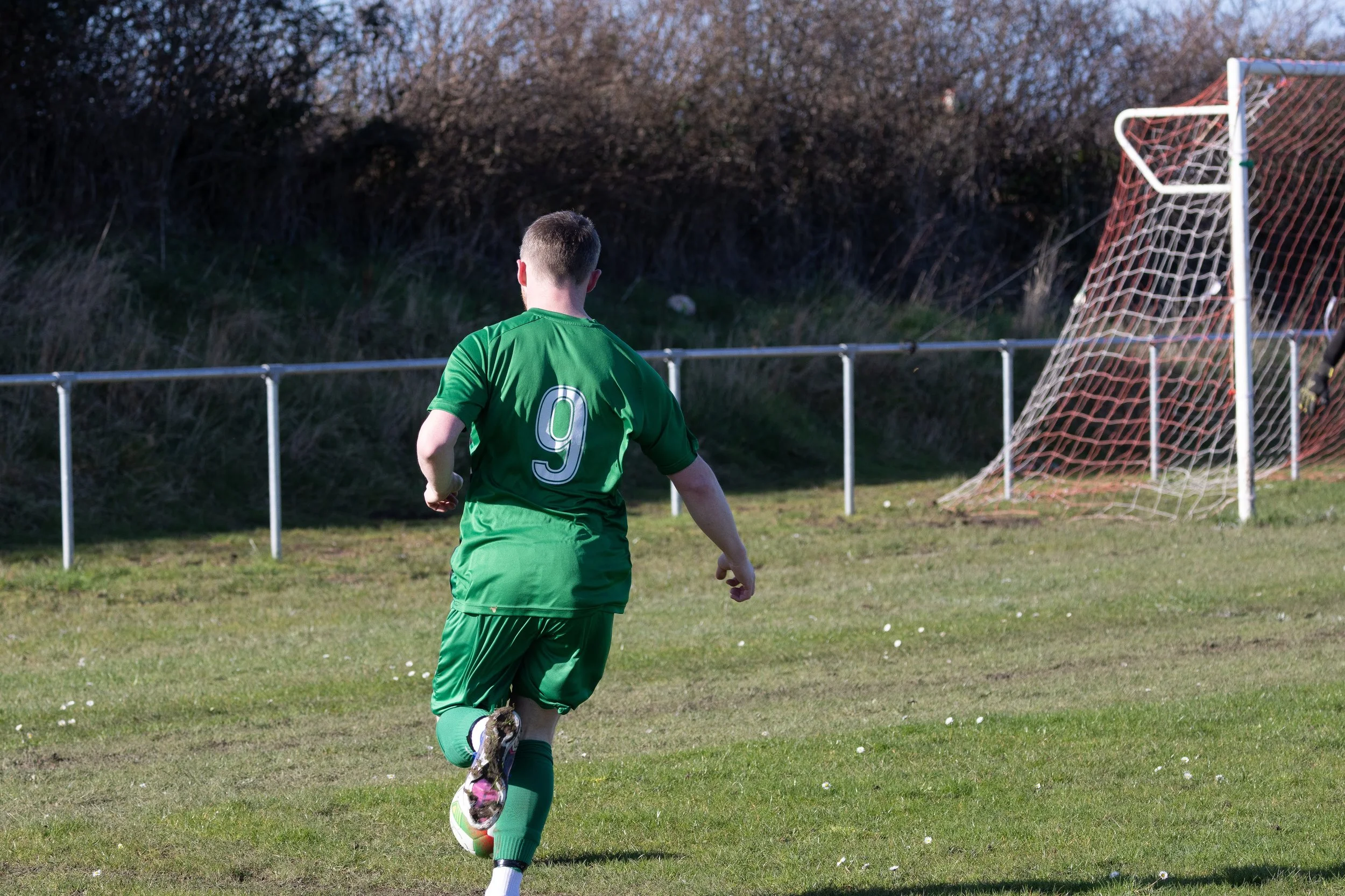 A young male soccer player in a green uniform with the number 9 on the back, running on a grassy field near a goalpost.