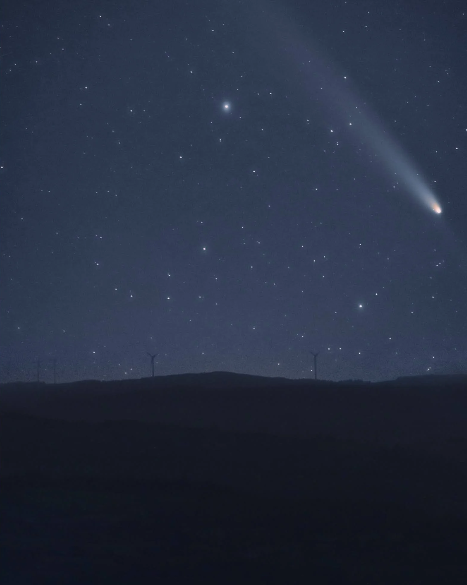 Night sky filled with stars and a bright comet with a visible tail, over a dark landscape with wind turbines.