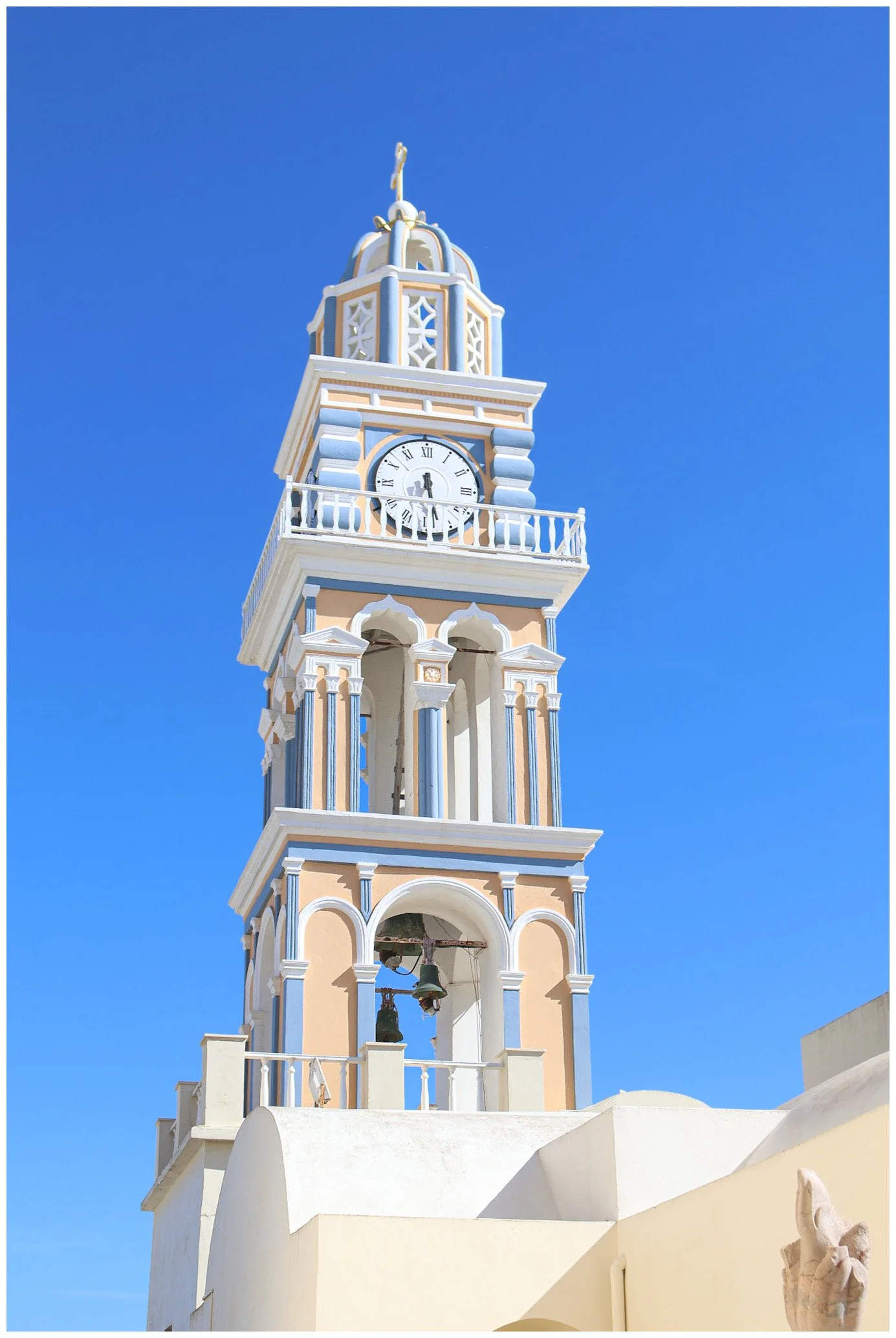 A tall clock tower with a beige and blue color scheme, featuring arched openings, decorative columns, and a clock face showing 1:53, set against a clear blue sky.