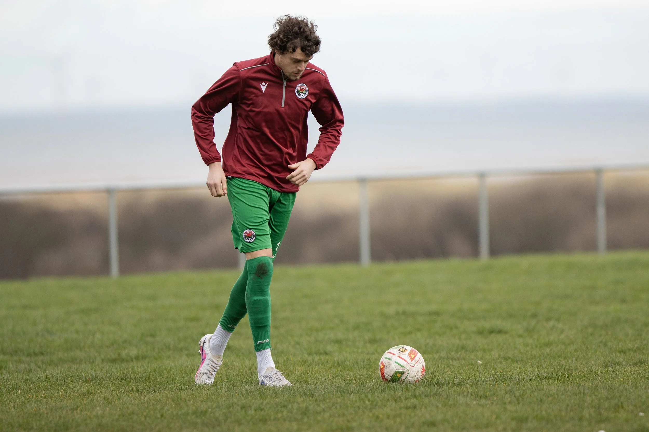 A soccer player in a red jacket and green shorts stands on a grassy field, looking down at a soccer ball.