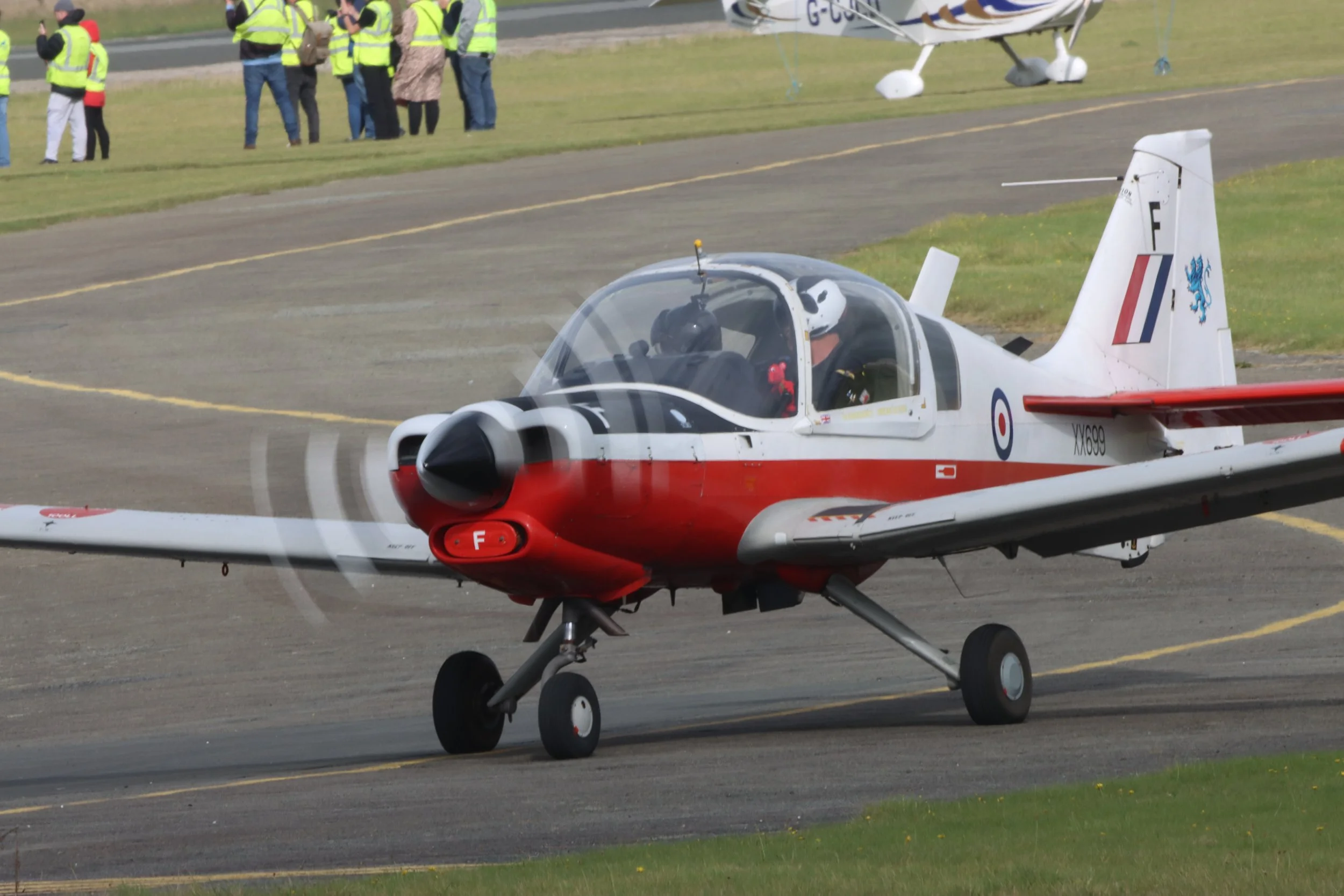 A red and white military aircraft taxiing on a runway with a pilot wearing a helmet inside. In the background, a group of onlookers in yellow safety vests and casual clothing stands near another small plane.