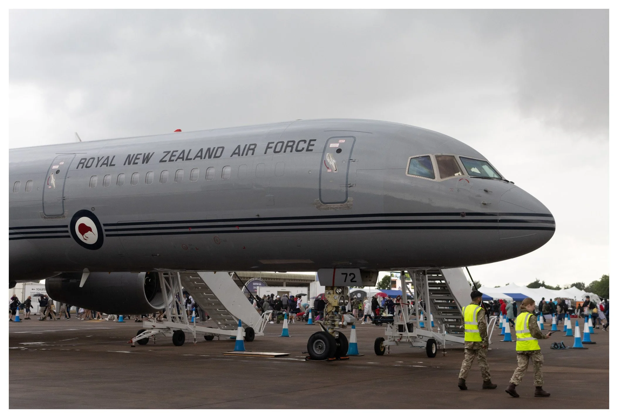 Military aircraft labeled 'Royal New Zealand Air Force' displayed on tarmac at an event, with people in the background and two personnel in yellow vests walking in front.