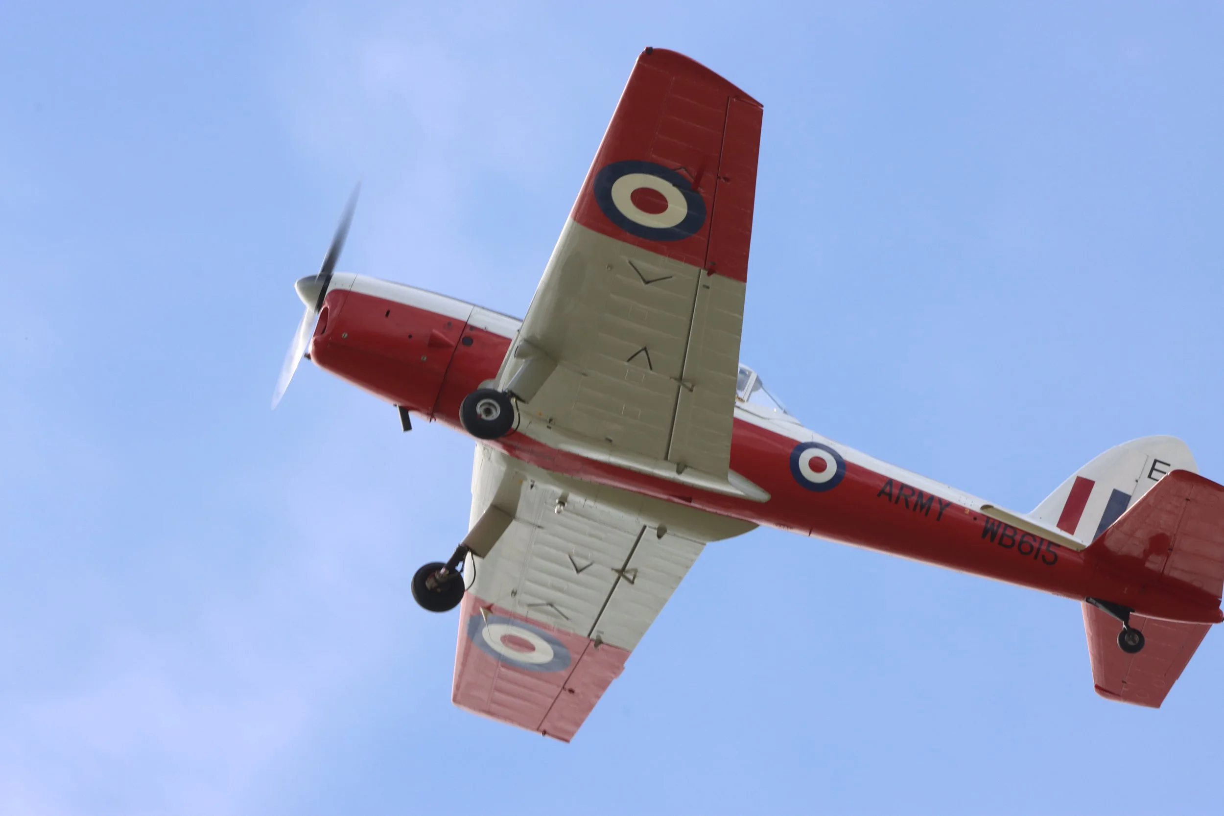 A small red and white vintage aircraft flying in a blue sky, with the words 'Army WB615' on its fuselage and roundel insignias on its wings and fuselage.