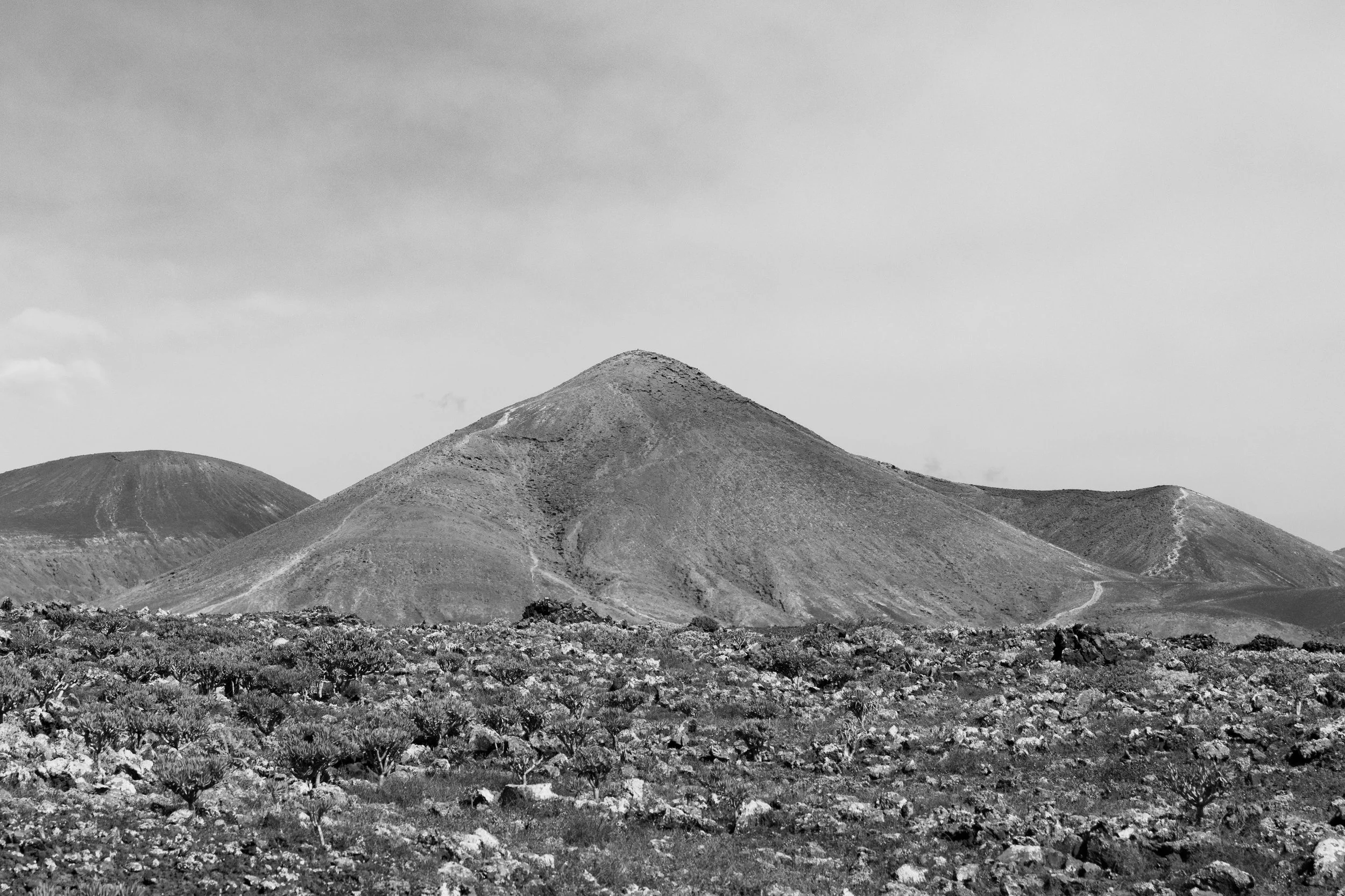 A black-and-white landscape featuring volcanic mountains with a rocky foreground and a cloudy sky.