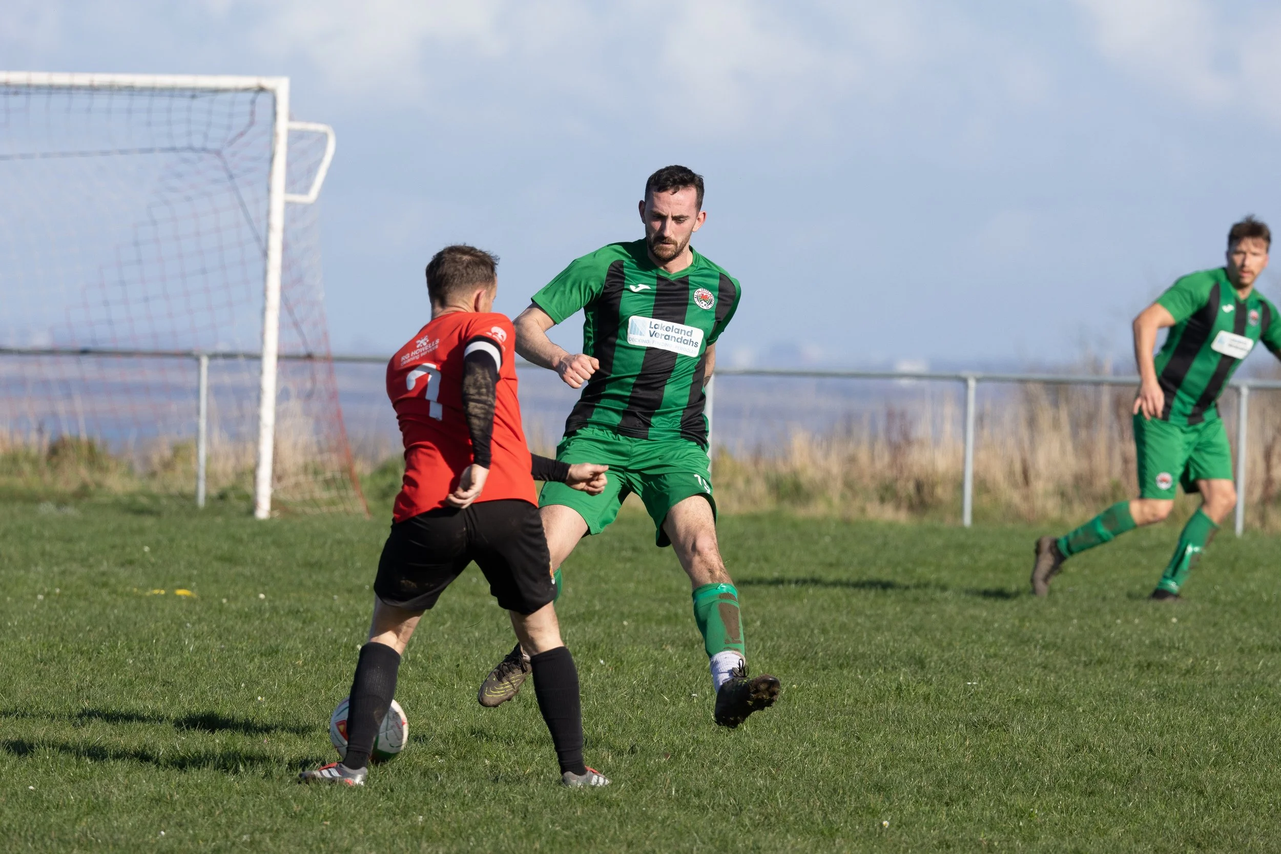 A soccer game with kids, one in a red uniform and two in green uniforms. The child in red is about to kick the ball while a tall player in green is trying to block or challenge him. The other player in green is running toward the play in the backgrou