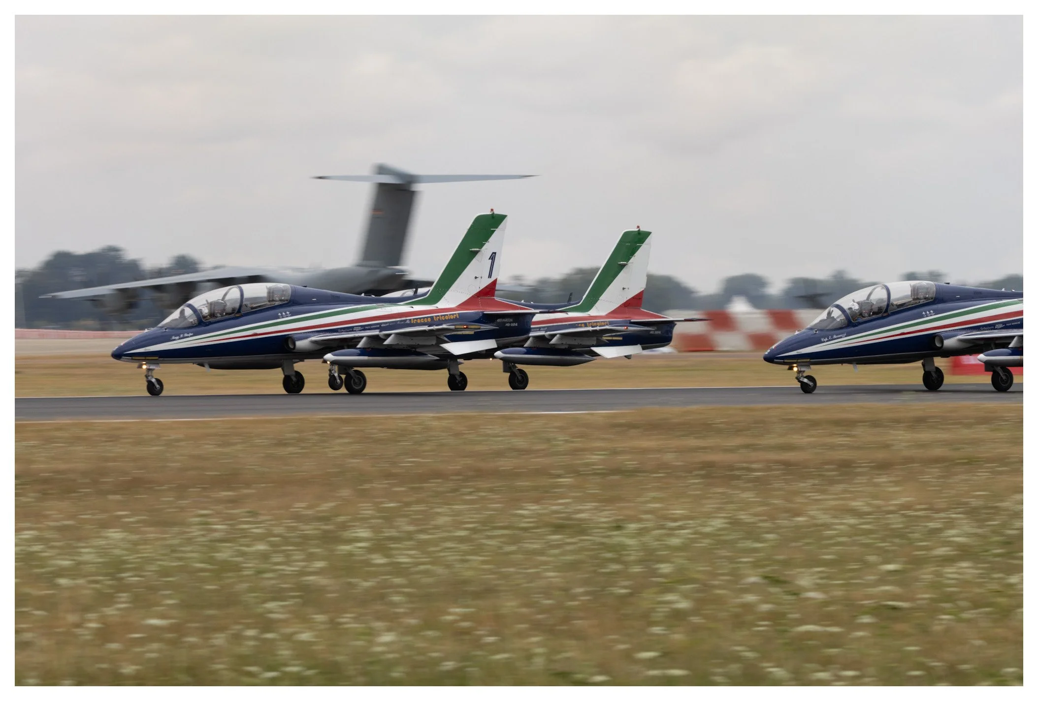 Two Italian fighter jets on the runway with a military transport aircraft taking off in the background under a cloudy sky.