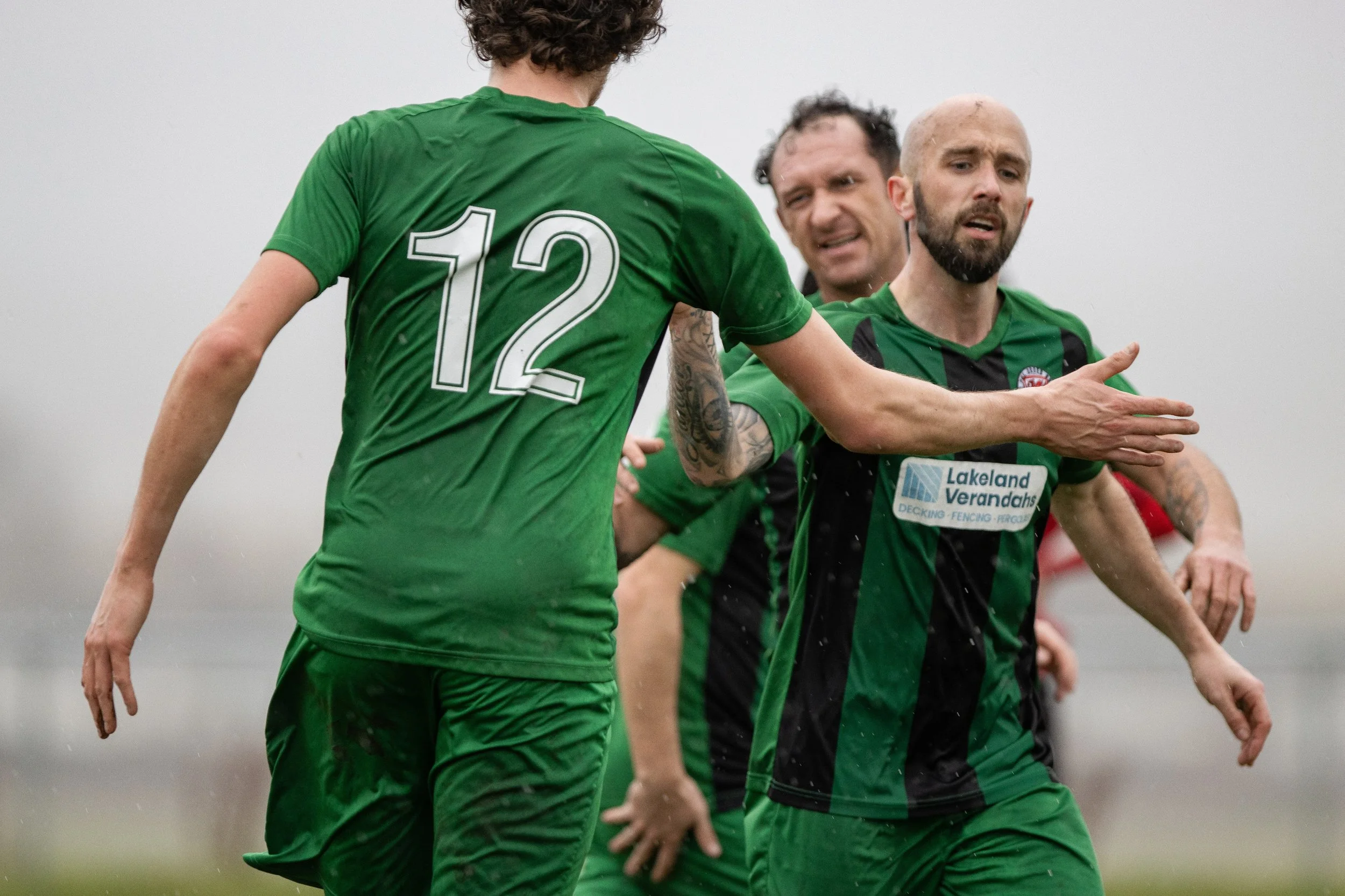 Three soccer players in green jerseys celebrating on the field during a rainy day. One player is extending his arm to high-five another with the number 12 on his back.