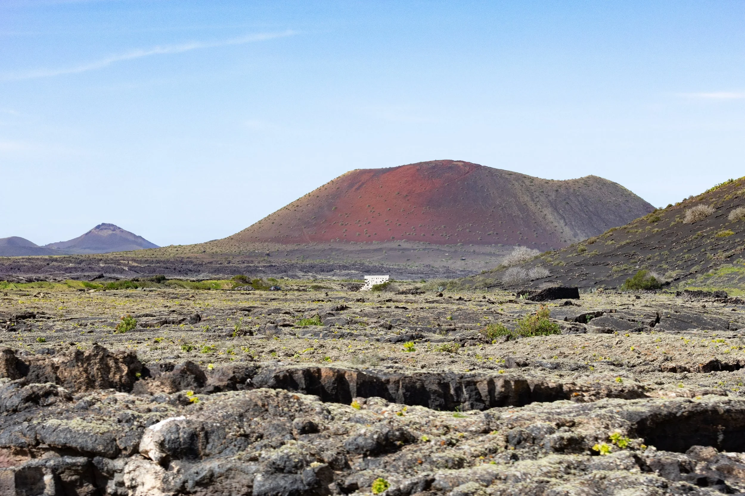 Volcanic landscape with a large reddish-brown volcano in the background, smaller volcanoes in the distance, and a rocky, barren foreground with sparse vegetation under a clear blue sky.