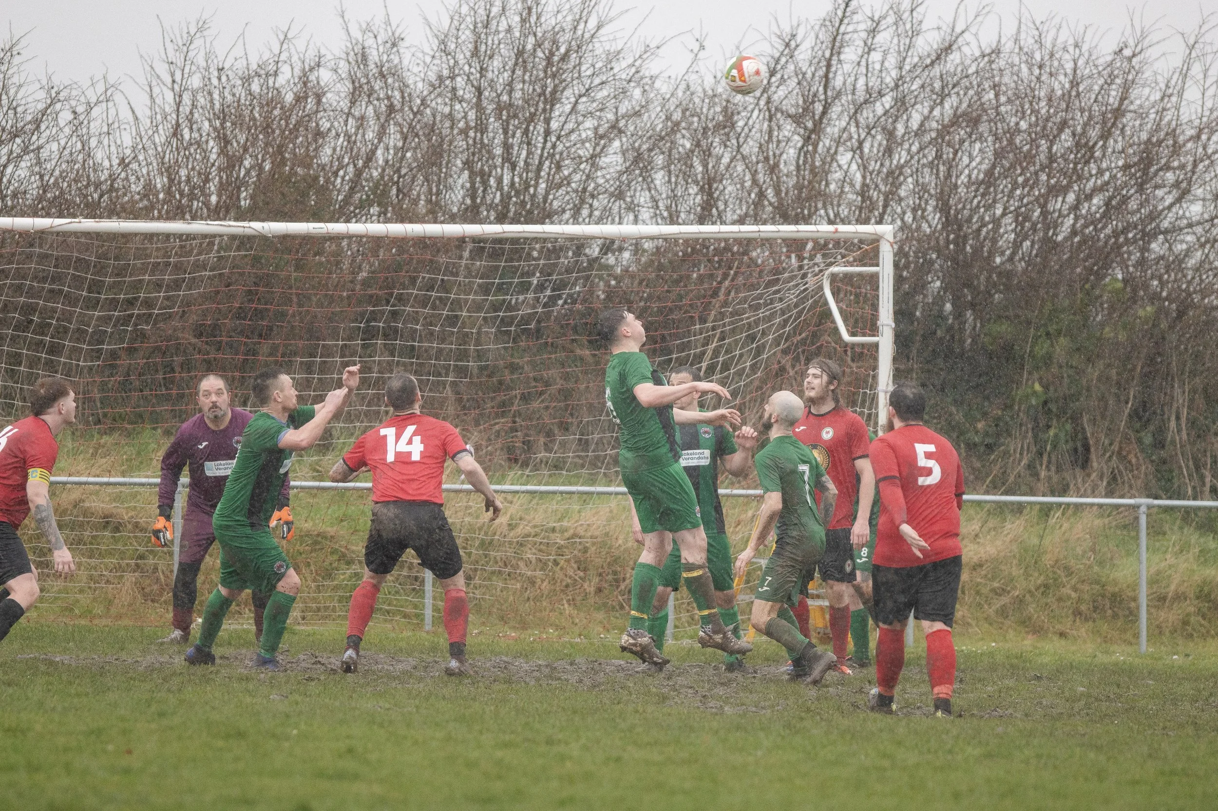 A group of soccer players in green and red uniforms jumping and competing for the ball in front of the goal on a rainy day.