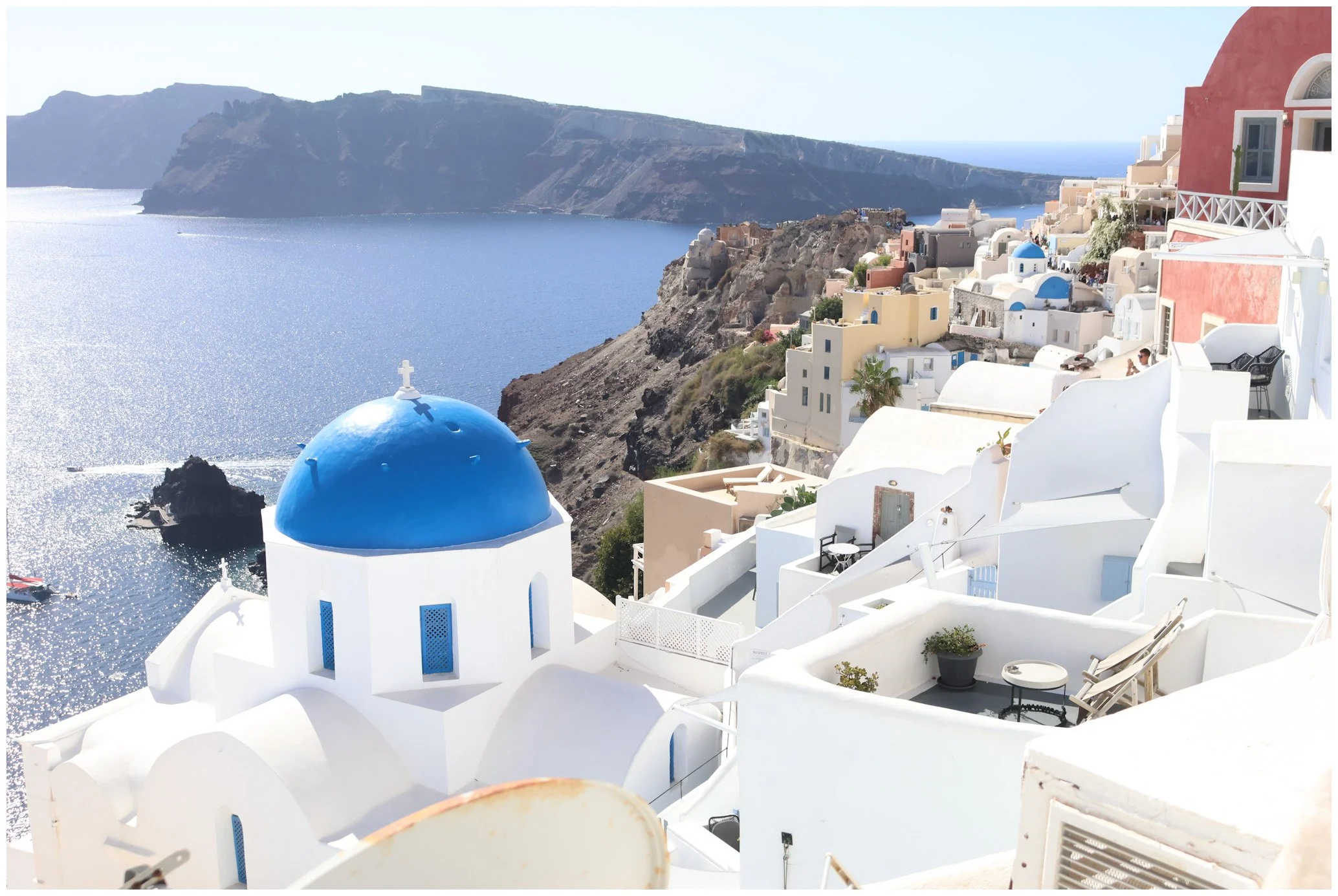 White buildings with blue domed roofs on a cliff overlooking the sea, with distant islands in the background.