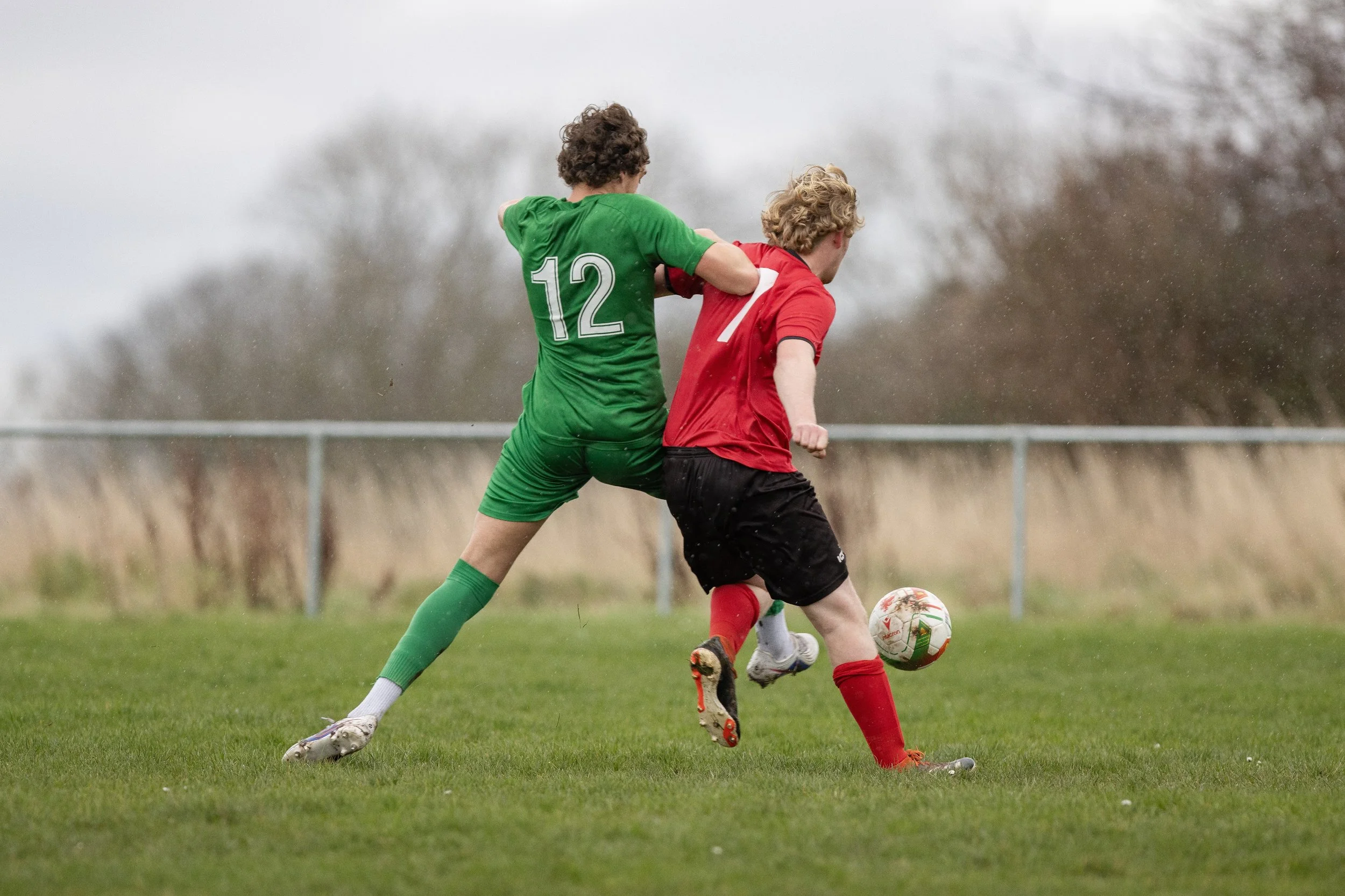 Two young boys playing soccer on a grassy field, one in a green uniform and the other in a red uniform, with the boy in red about to kick the ball.