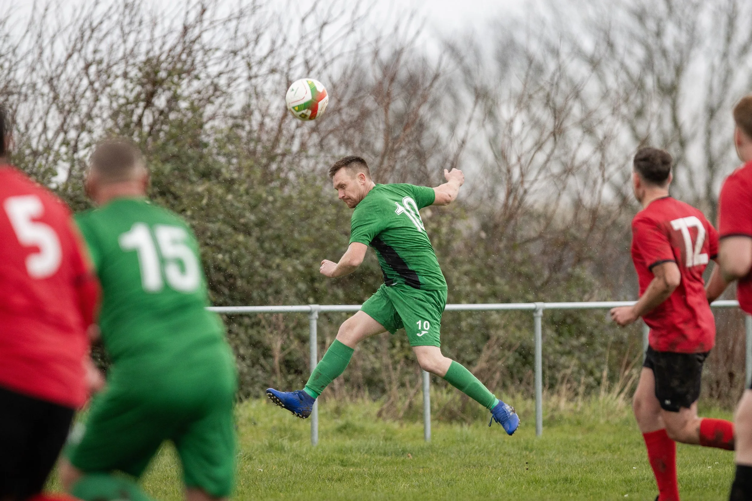 A soccer player in green uniform jumping to head the ball during a match with opponents in red uniforms nearby.