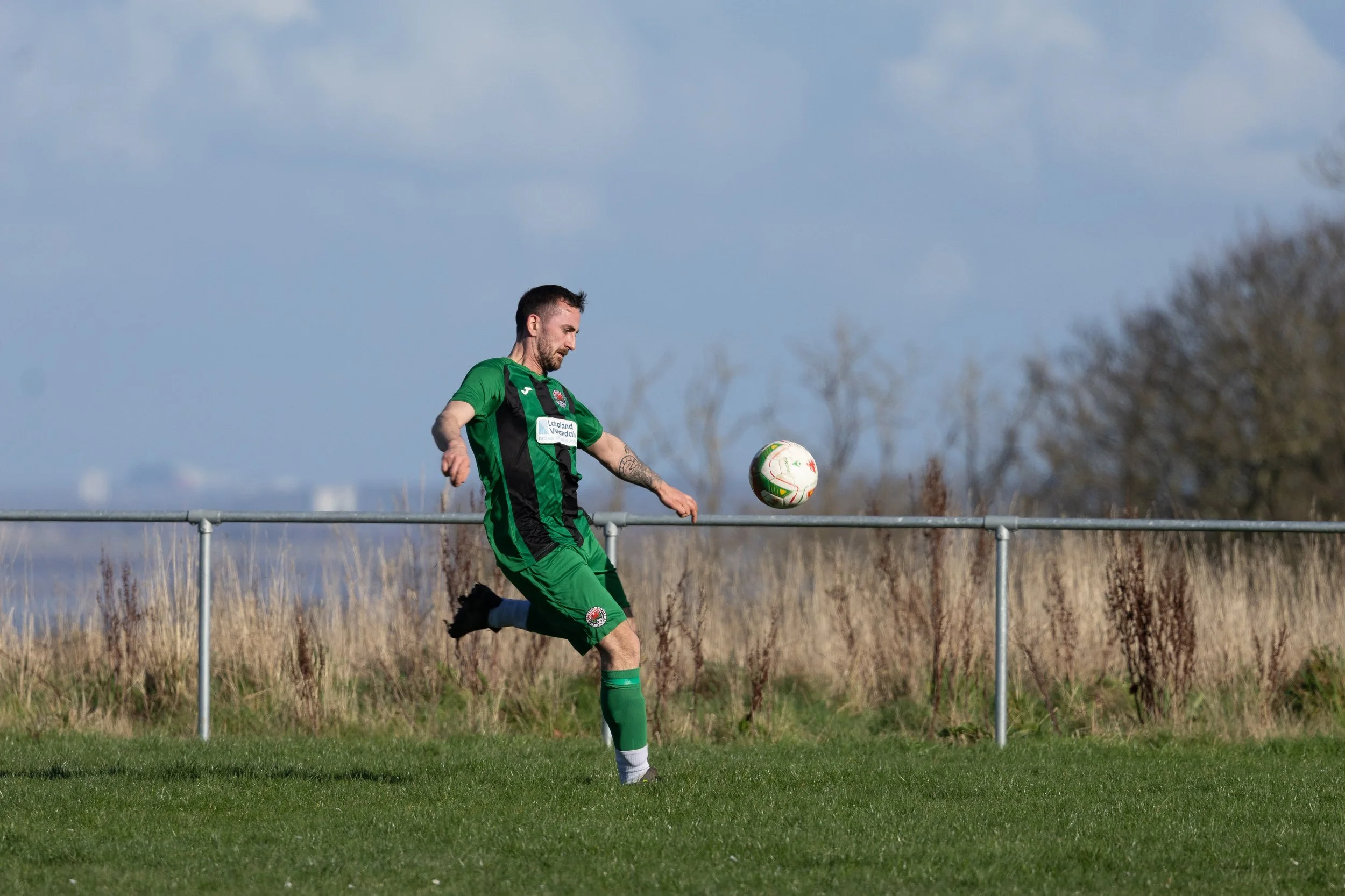 A soccer player in a green and black uniform kicking a ball on a grass field during daytime.