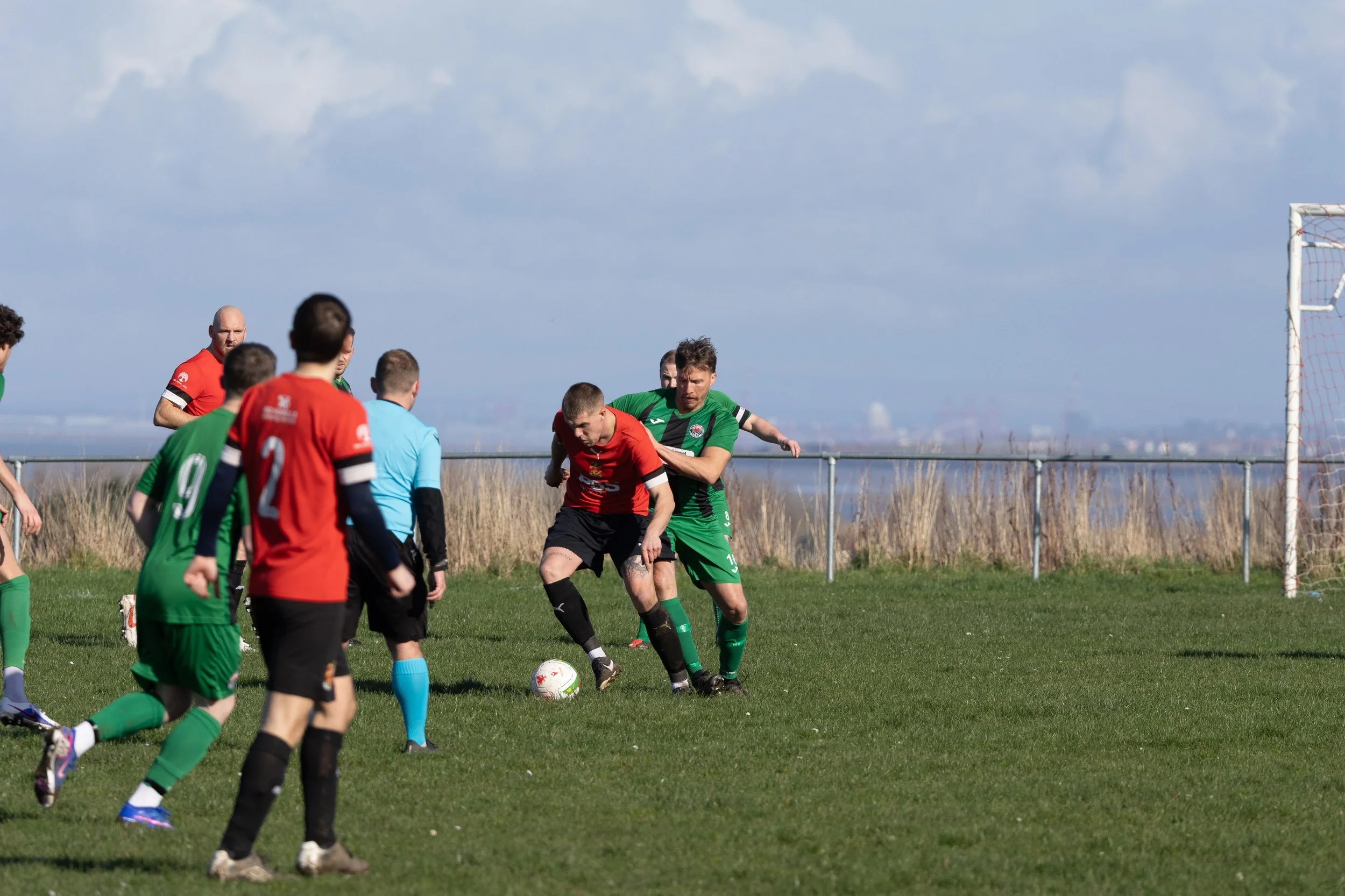 Soccer match with players in red and green jerseys competing for the ball on a grassy field, with a blue sky and water in the background.