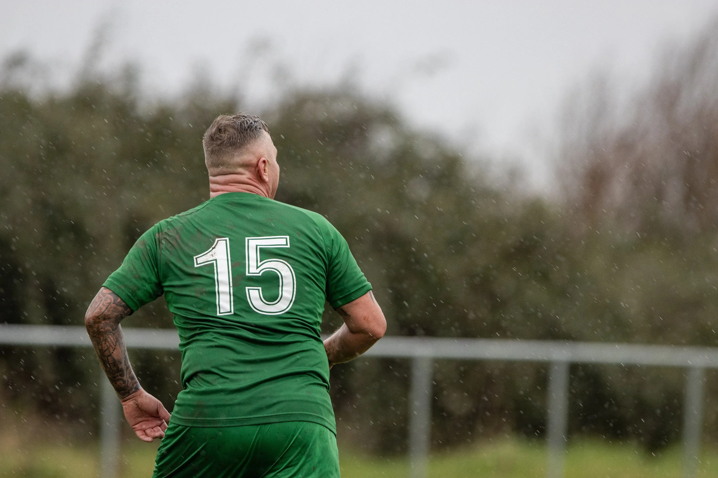 A soccer player wearing a green jersey with the number 15 on the back, standing outdoors on a rainy day with trees and a fence in the background.