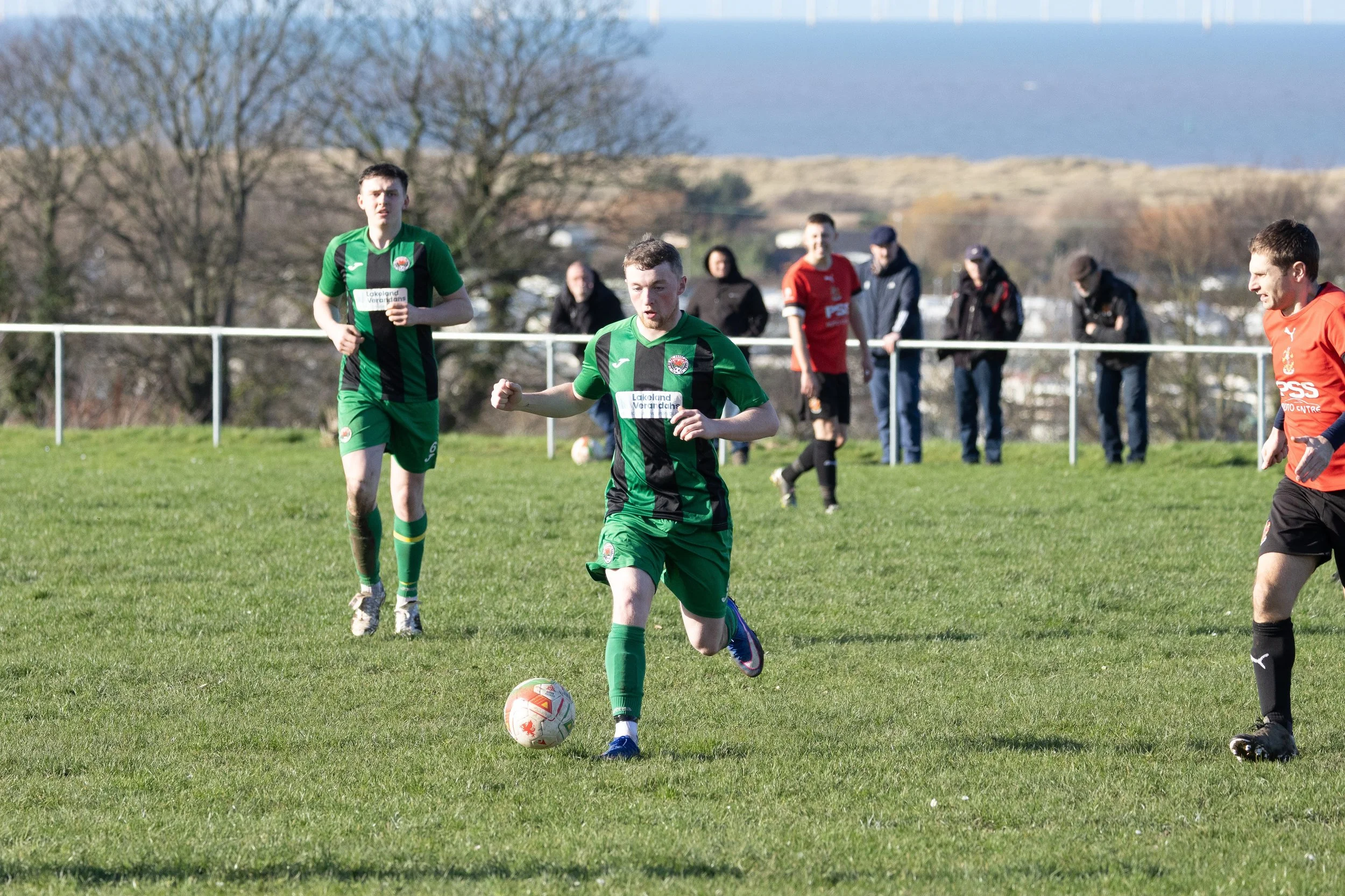 A soccer player in a green uniform is running with the ball on a grassy field, with other players and spectators in the background.