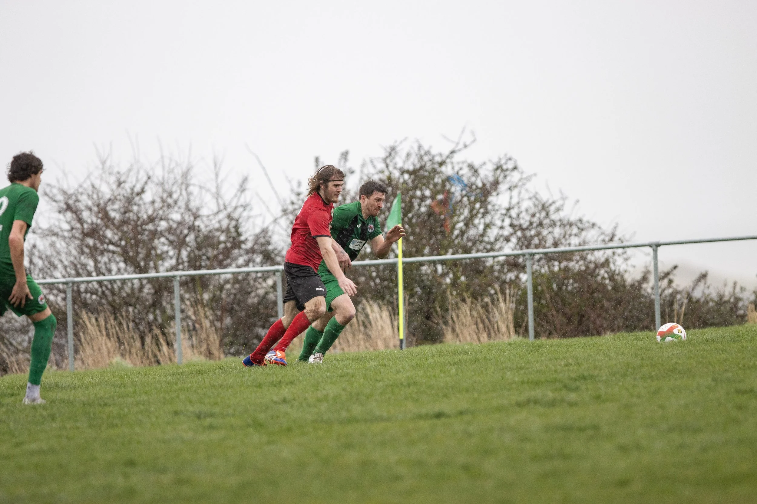 Two soccer players, one in a red jersey and the other in a green jersey, competing for the ball near the corner flag on a grass field with a cloudy sky and trees in the background.