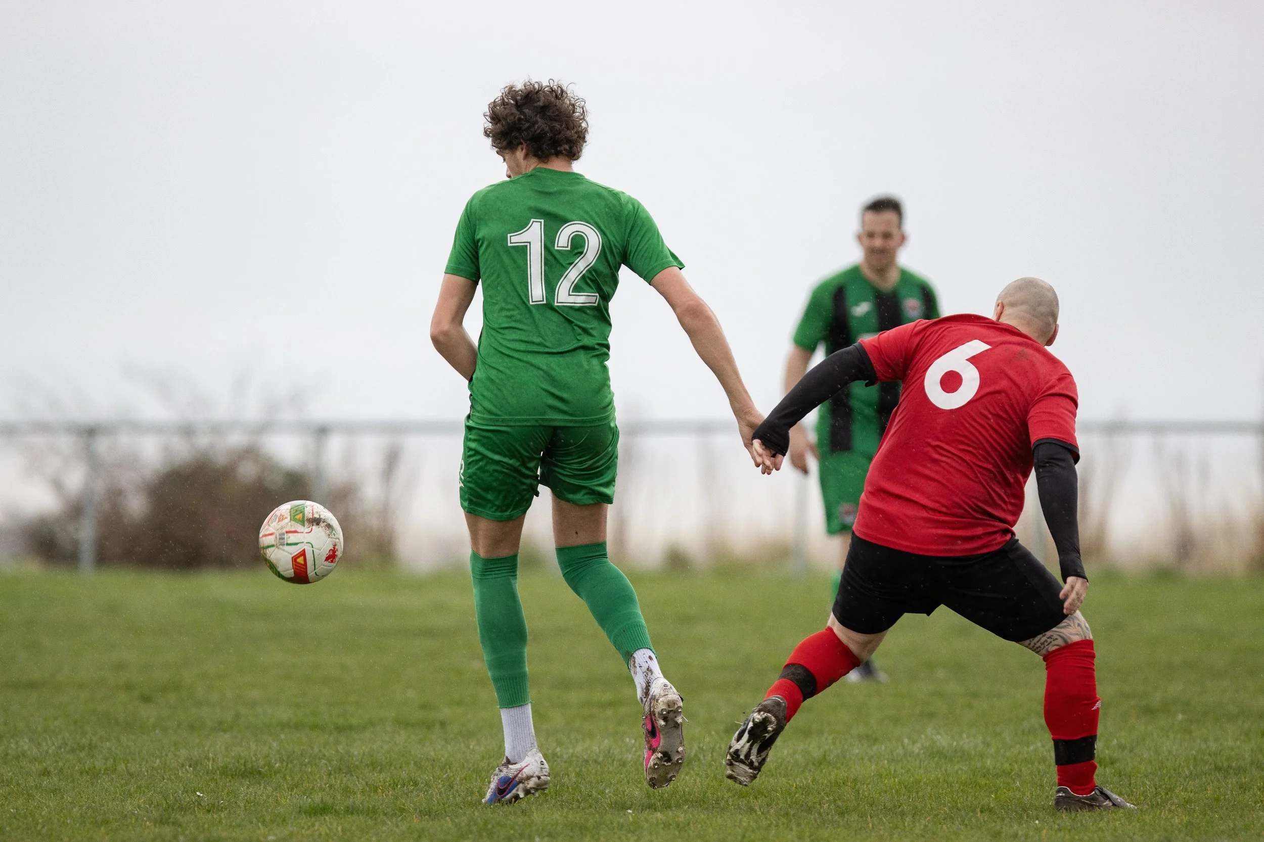 Soccer players in green and red uniforms holding hands on the field, with a soccer ball nearby, during a game.
