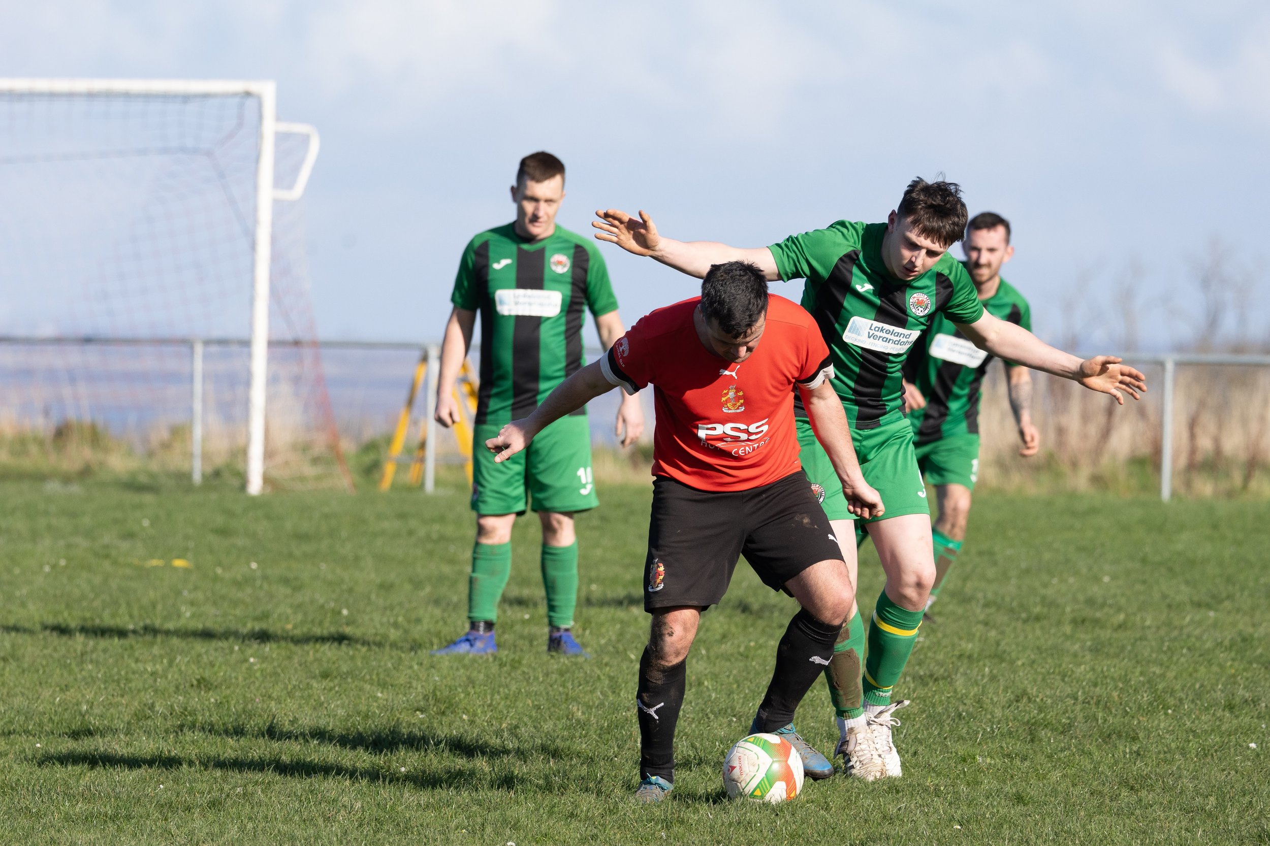 A soccer game with four male players on a grassy field, one in a red uniform controlling the ball while three in green uniforms watch. A goal net is visible in the background.