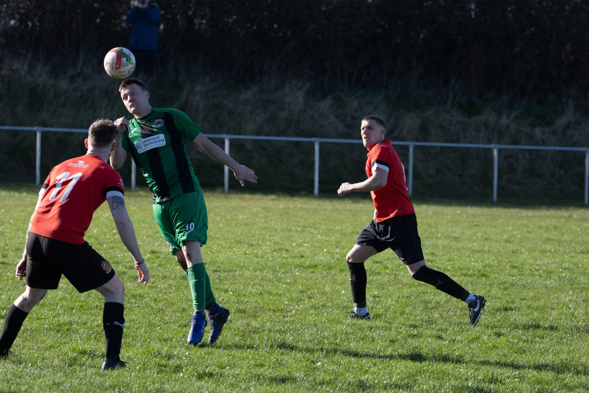 Soccer players in green and red jerseys competing for the ball on a grassy field in sunlight.