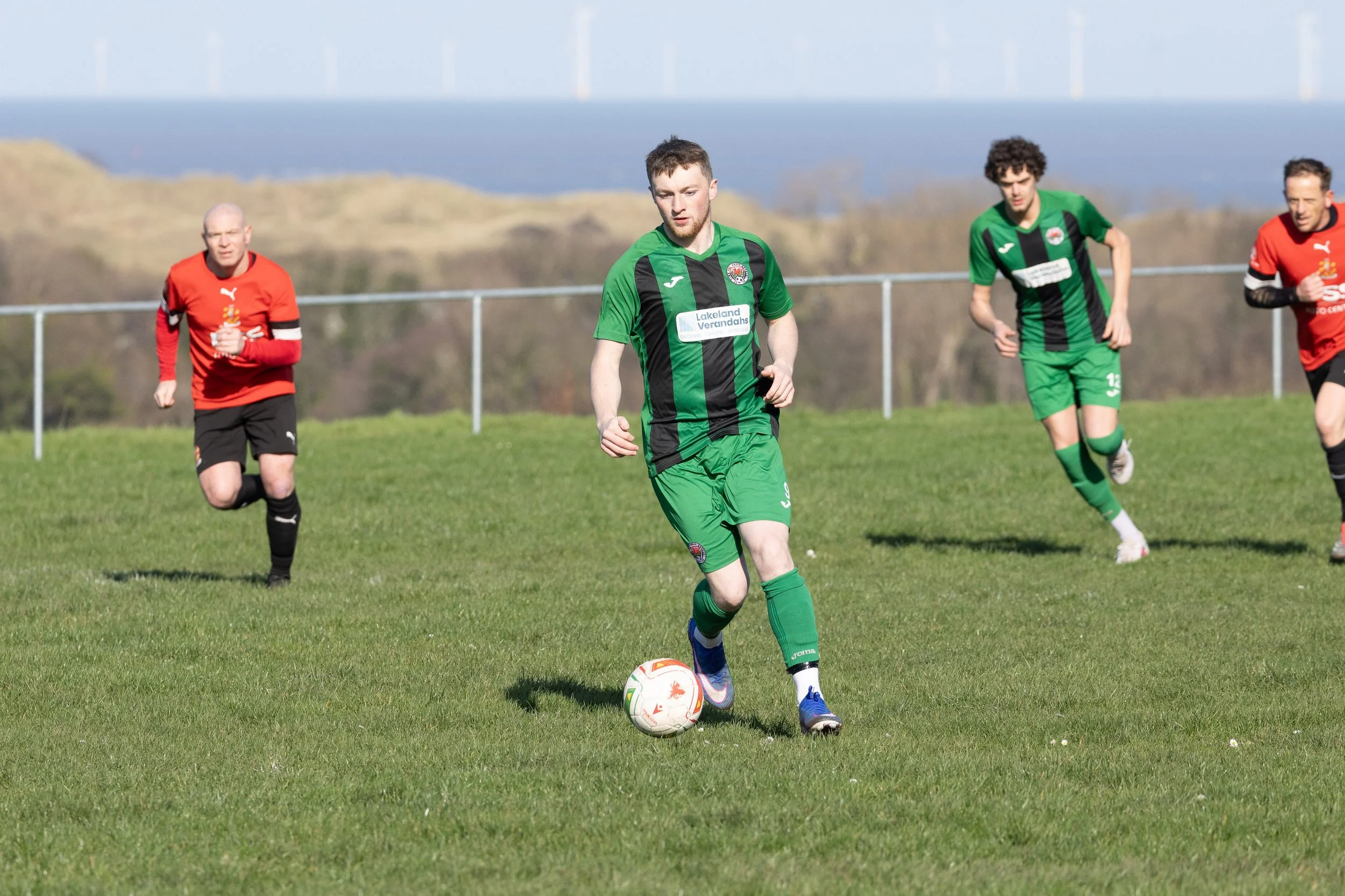 Soccer players on a field during a match, with one player in green and black about to kick the ball, while others run nearby.