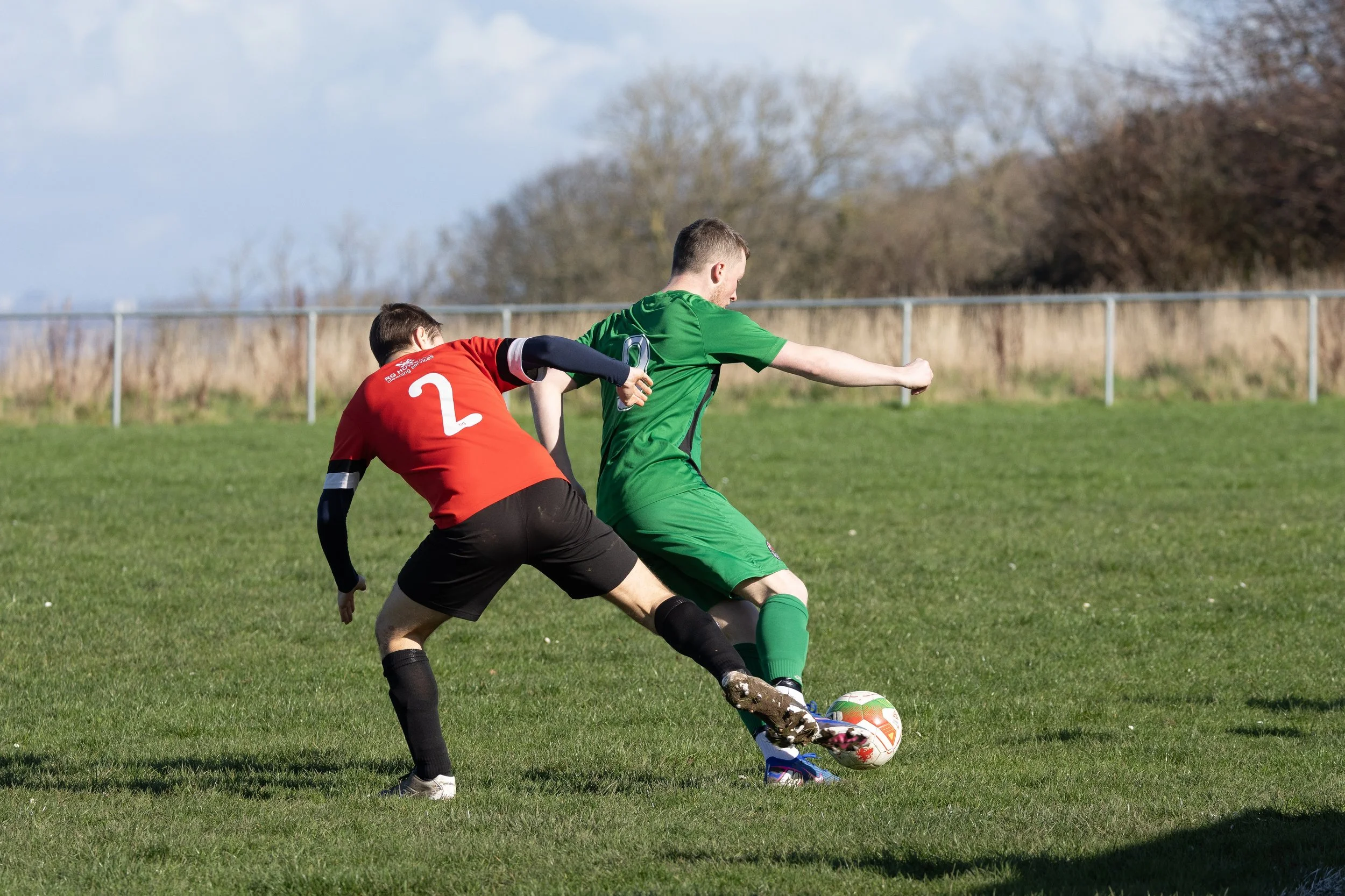 Two soccer players compete for the ball on a grassy field during daytime; one player wears a red jersey with the number 2, and the other wears a green uniform.