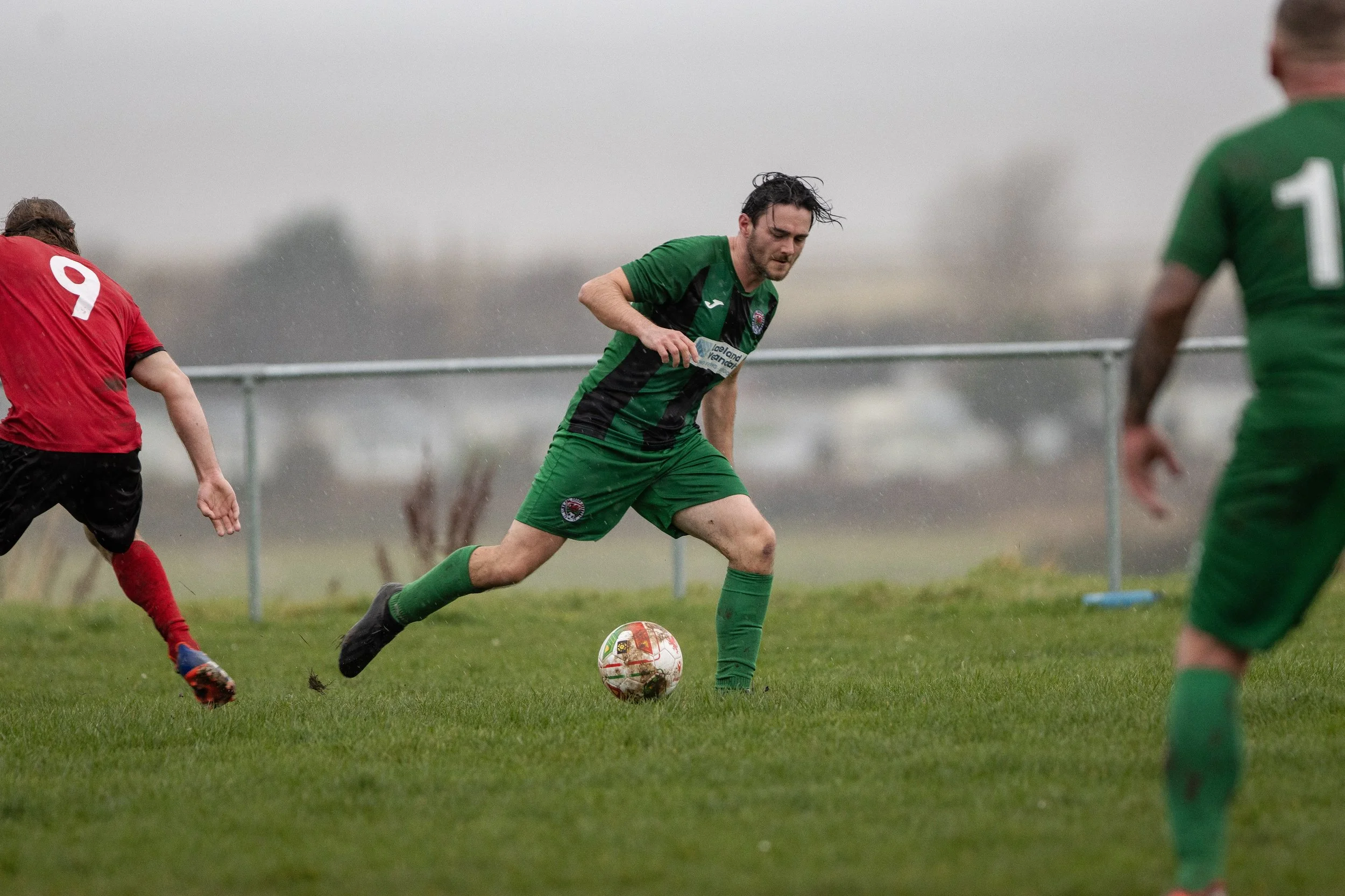 A soccer match with players in green and red jerseys playing on a grassy field during rainy weather.
