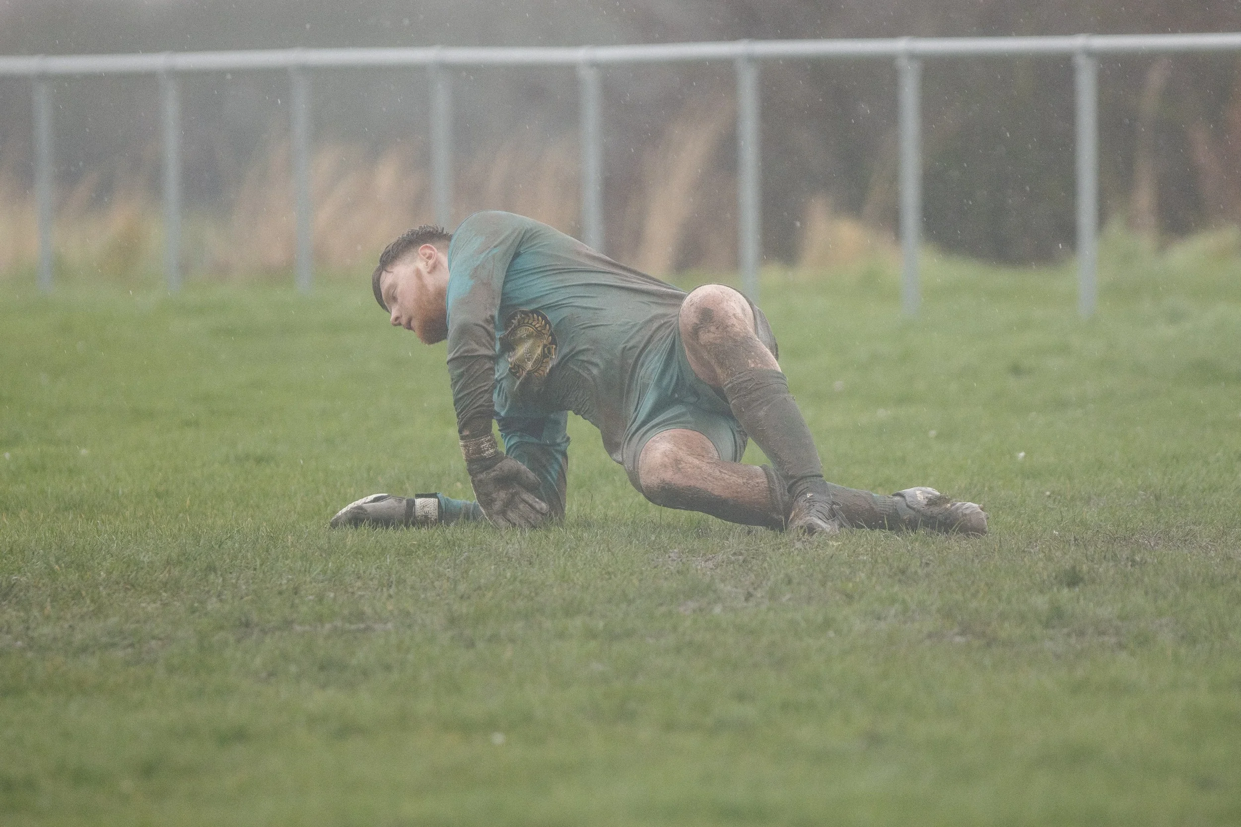 Soccer player kneeling on muddy field, exhausted and muddy, wearing a green uniform, in rainy weather.