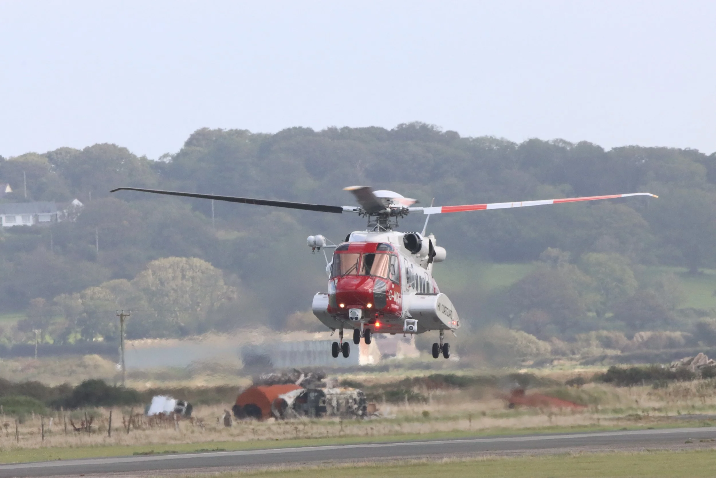 A rescue helicopter hovering above a crashed vehicle on an open field with green hills in the background.