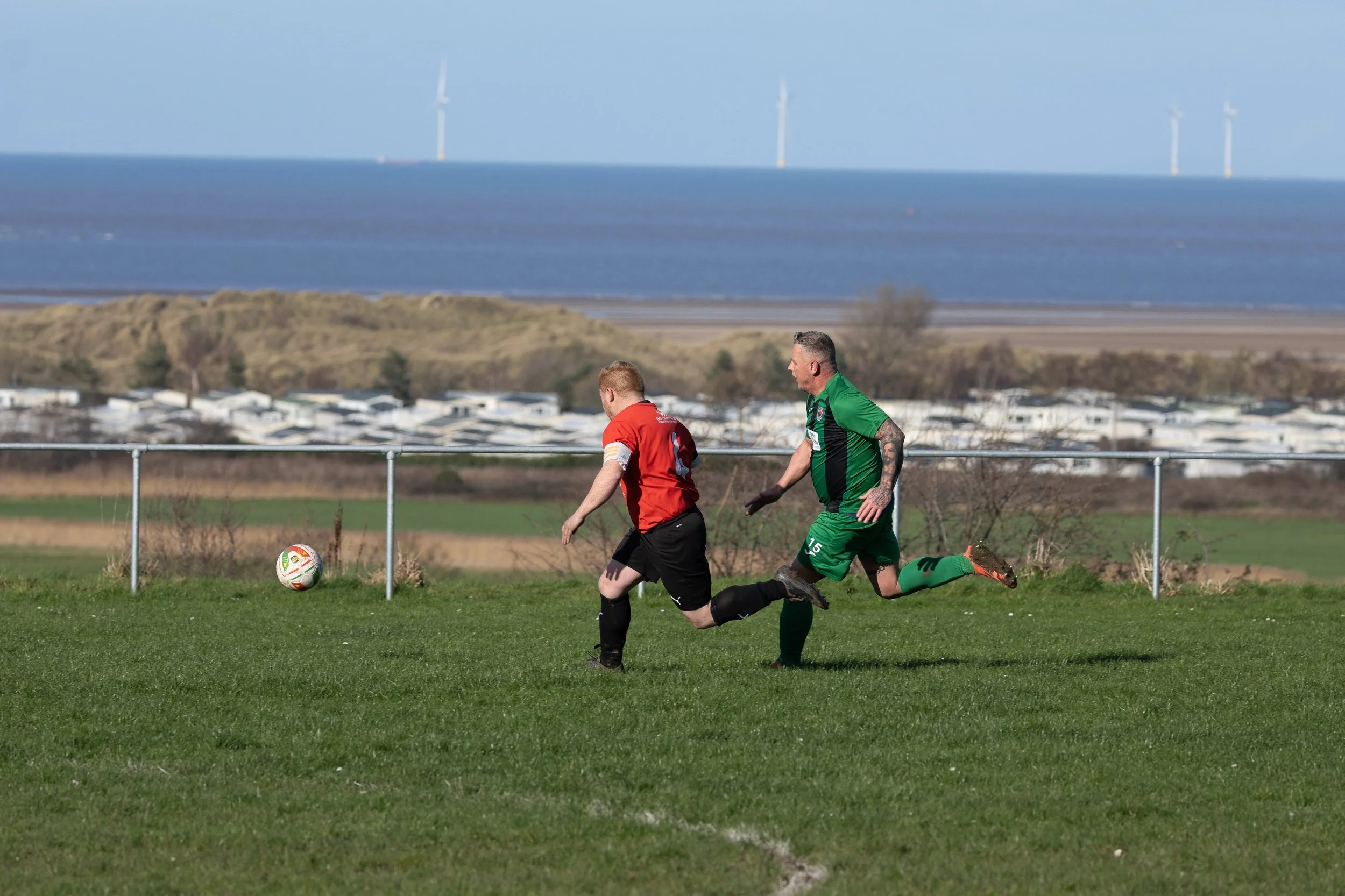 Two soccer players, one in red and one in green, chase a soccer ball on a field with a barrier fence and a scenic backdrop of water and wind turbines.
