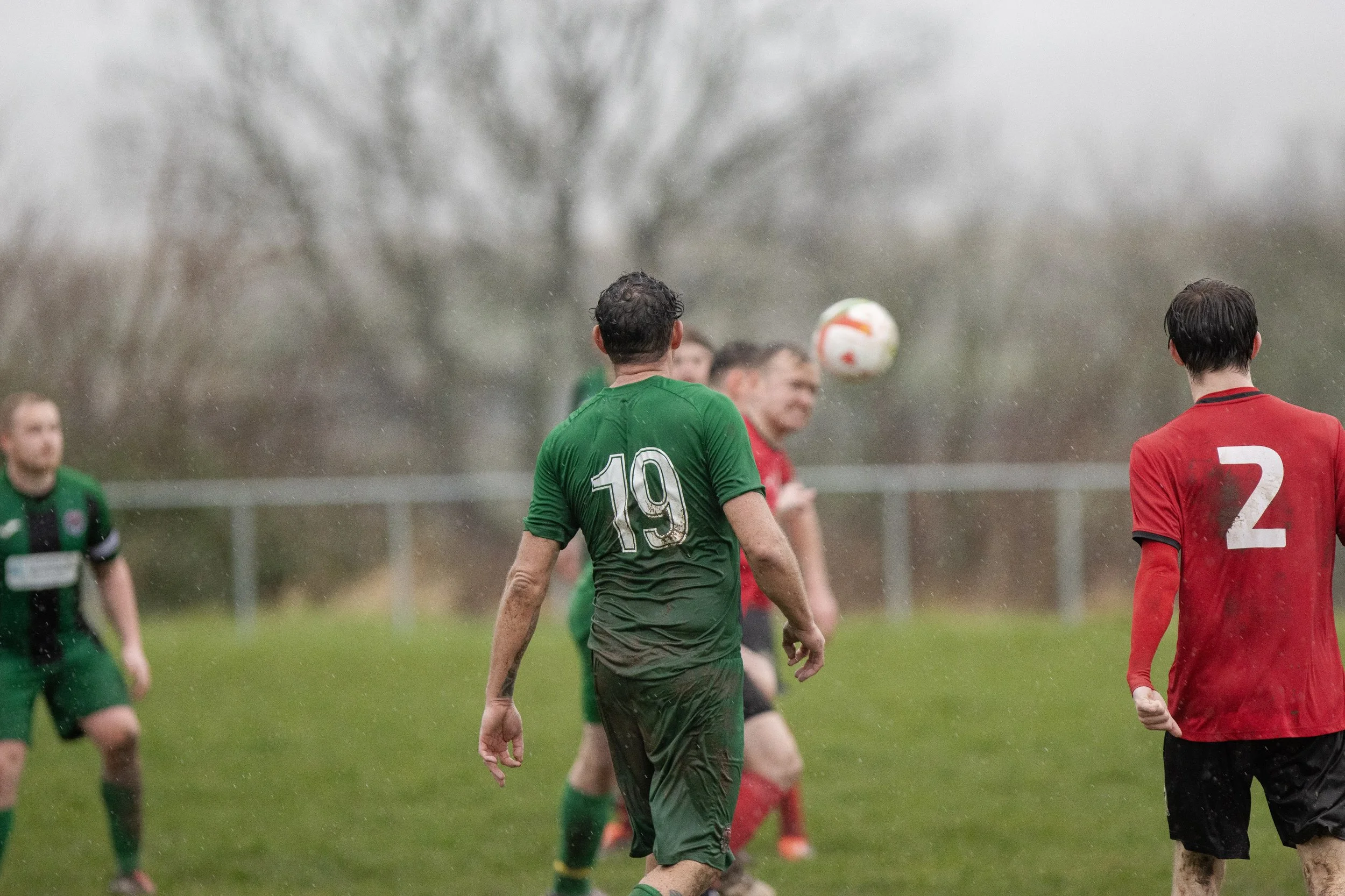 Mens soccer match in the rain with players wearing green and red jerseys, on a grass field with trees in the background.