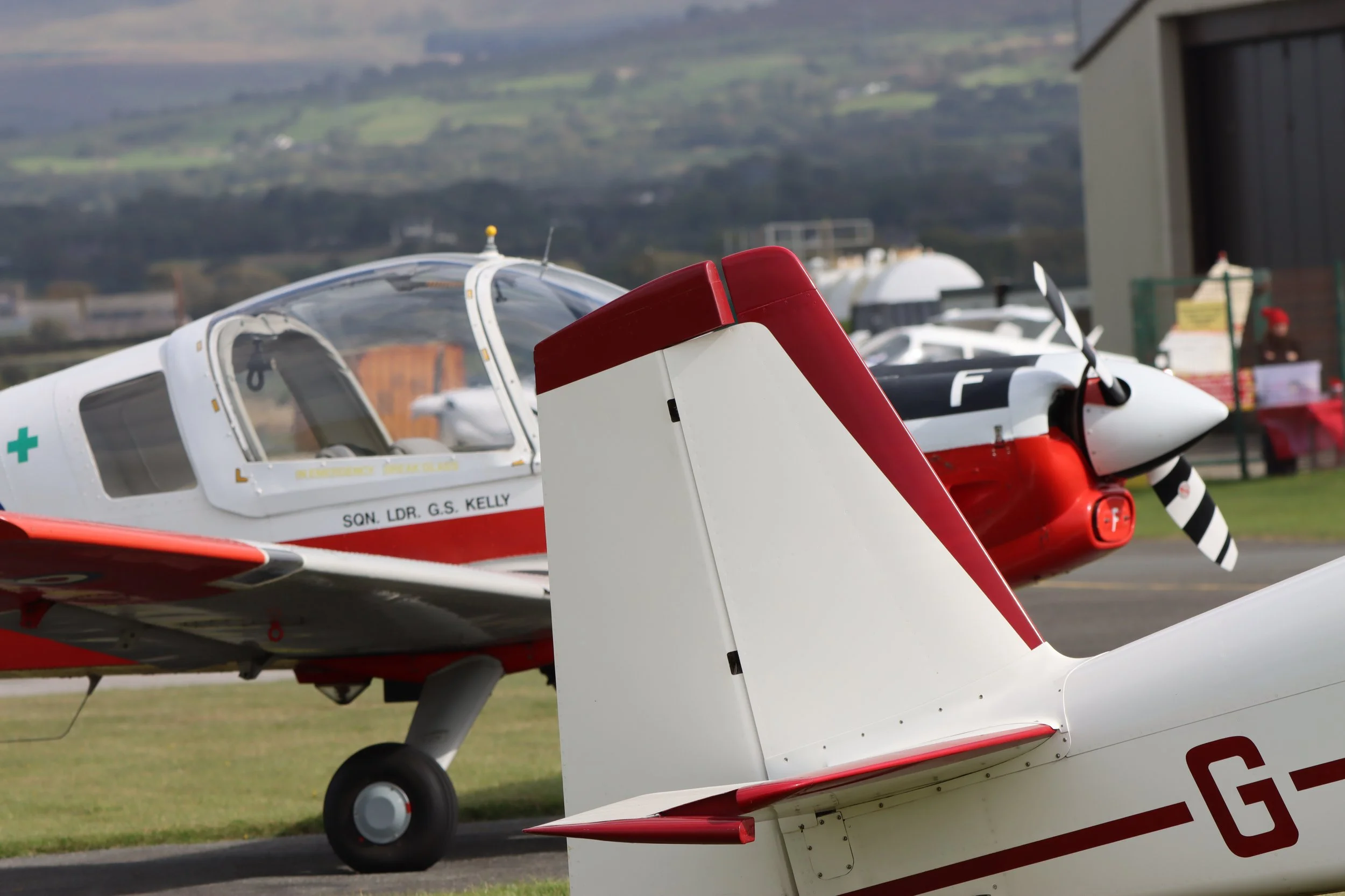 Two small white training aircraft with red accents parked on a grass airstrip, with mountains in the background.