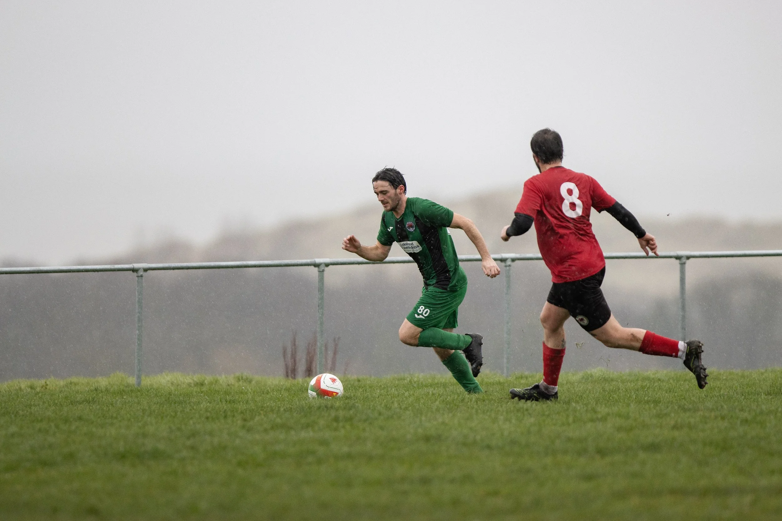 Two soccer players competing for the ball on a grassy field on a rainy day, with overcast sky in the background.