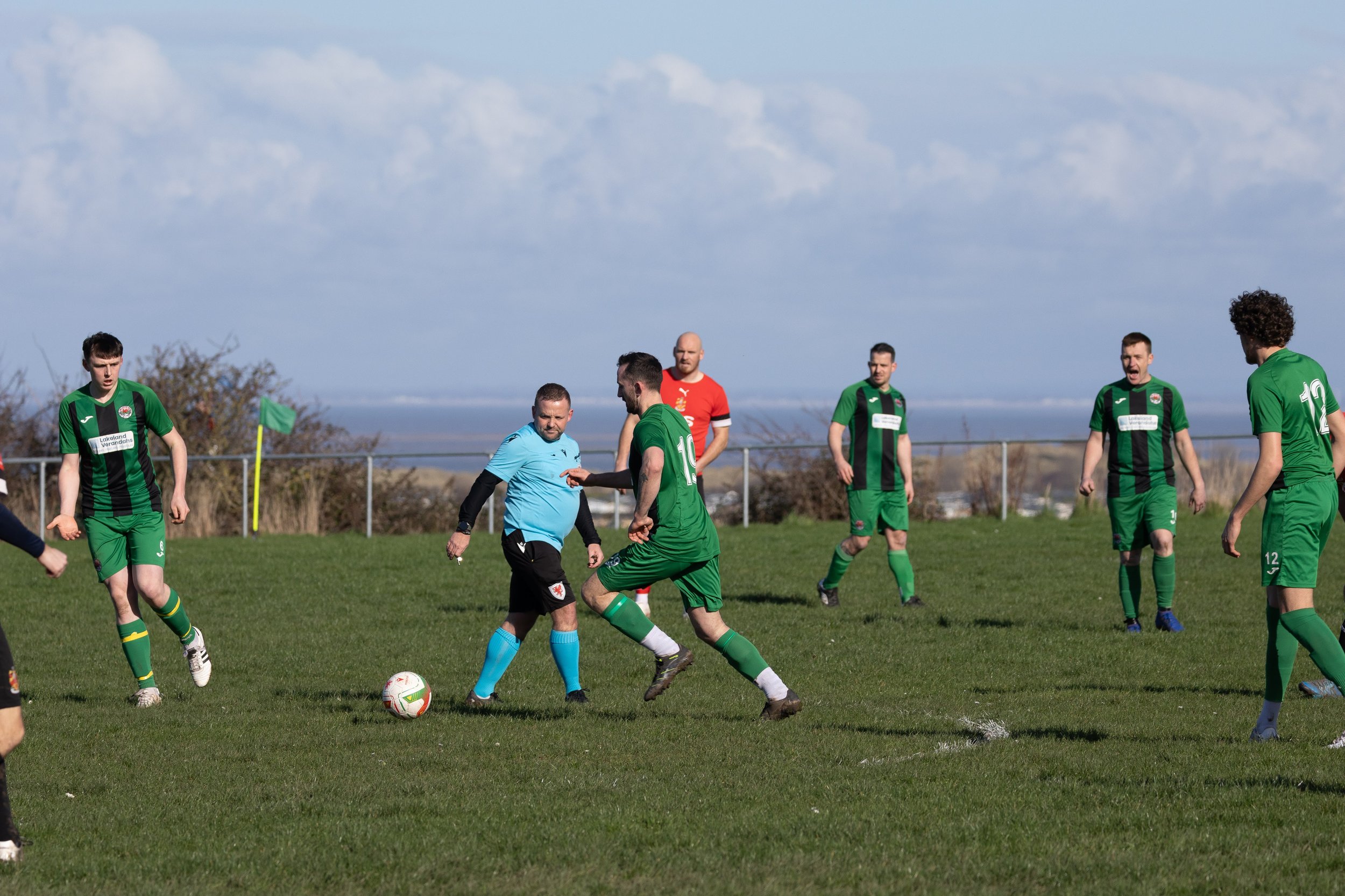 A soccer match with players in green and black jerseys, along with referees, on a grass field under a partly cloudy sky, with a distant landscape in the background.