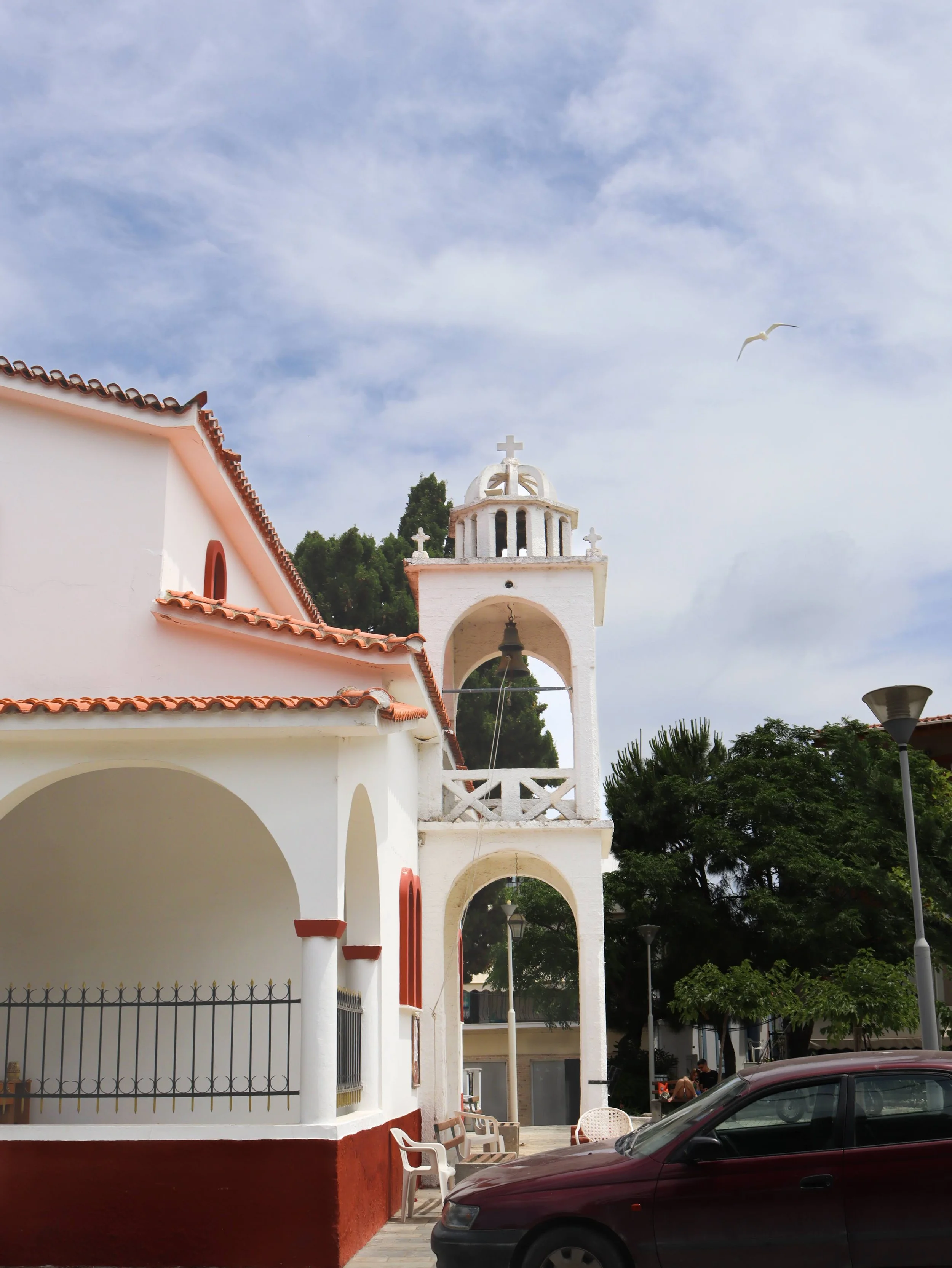A white church with red accents, a bell tower with a cross on top, and a partly cloudy sky. A maroon car is parked in front, and trees are visible in the background.