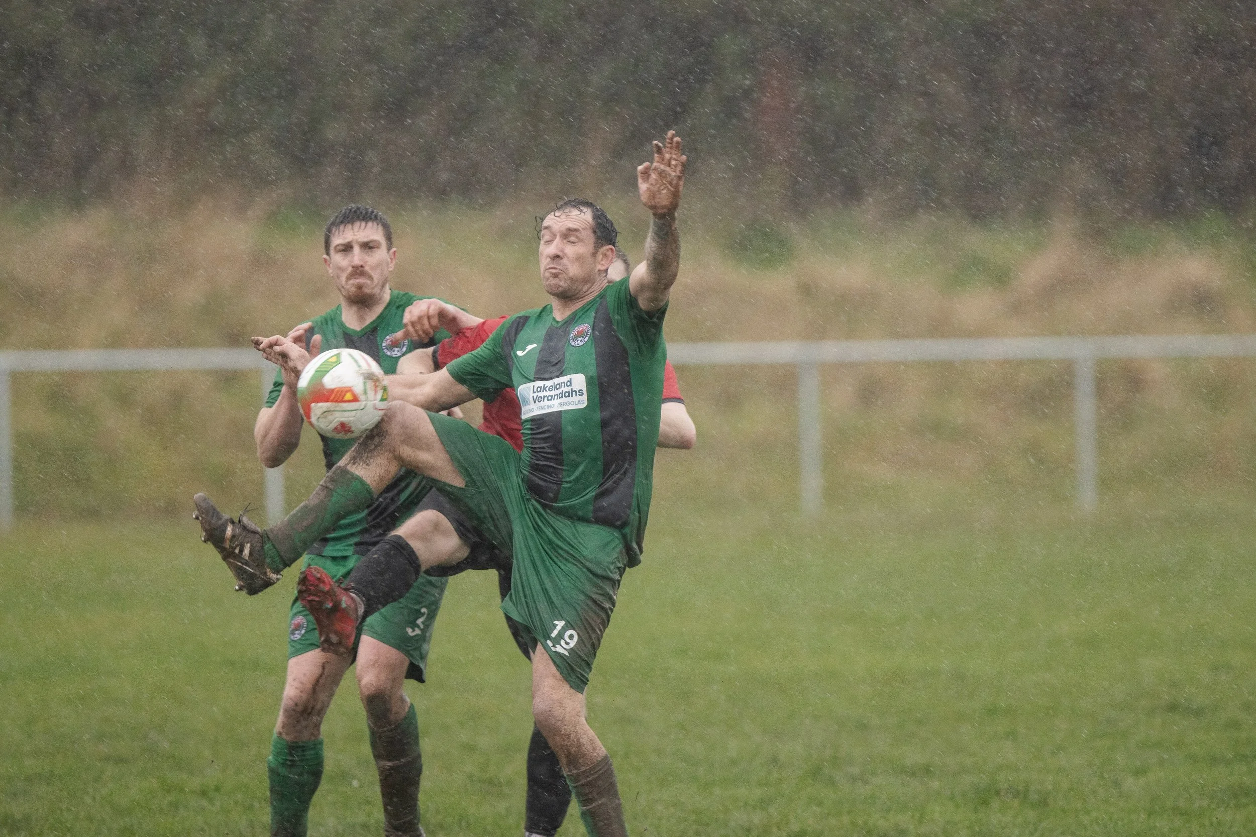 Two soccer players in green and black jerseys compete for the ball in a rain-soaked field, with a fence and blurred outdoor background.