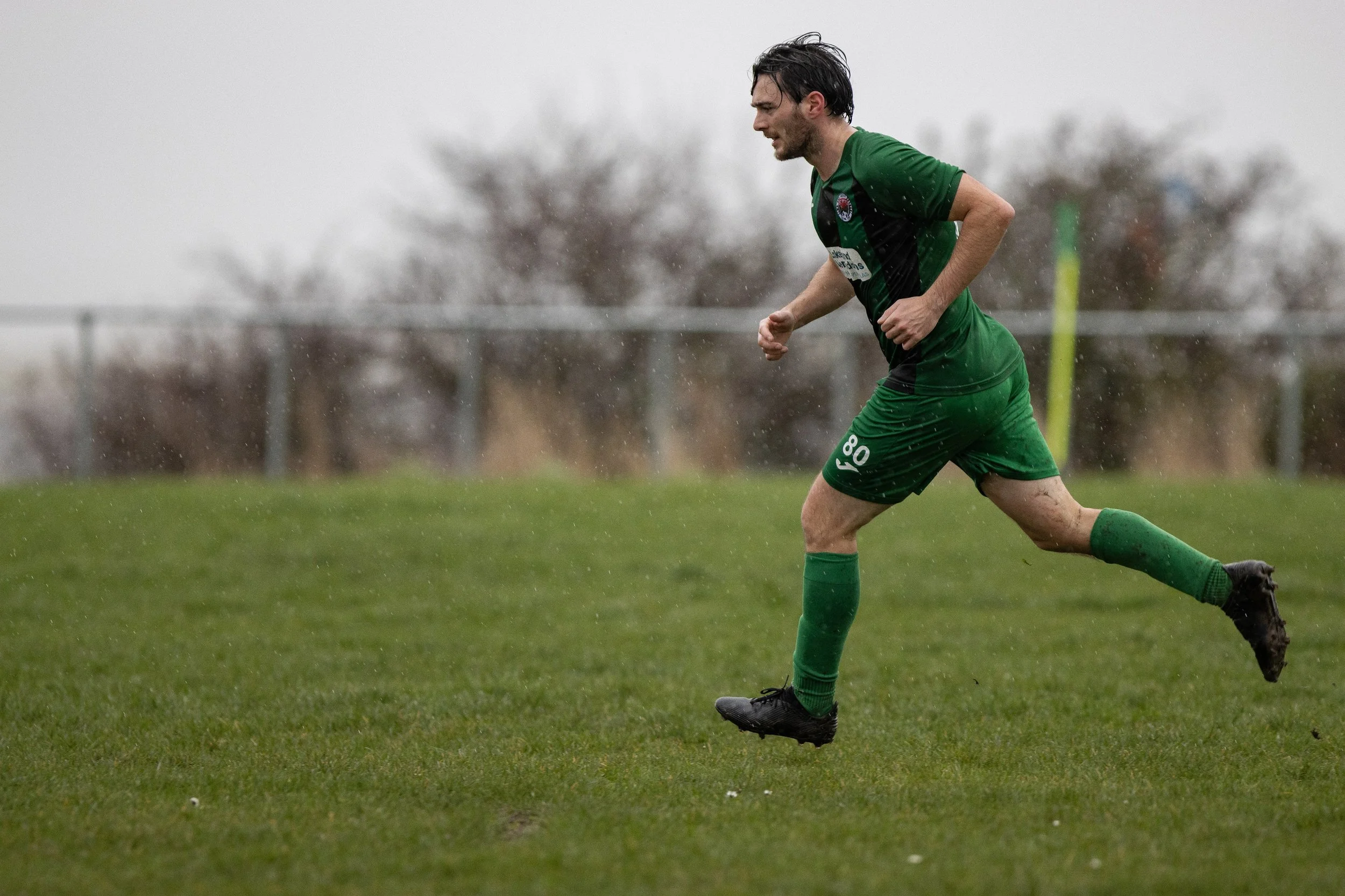 A male soccer player running on a wet field during a game, wearing a green jersey and shorts, with black cleats, under overcast skies and rainy weather.