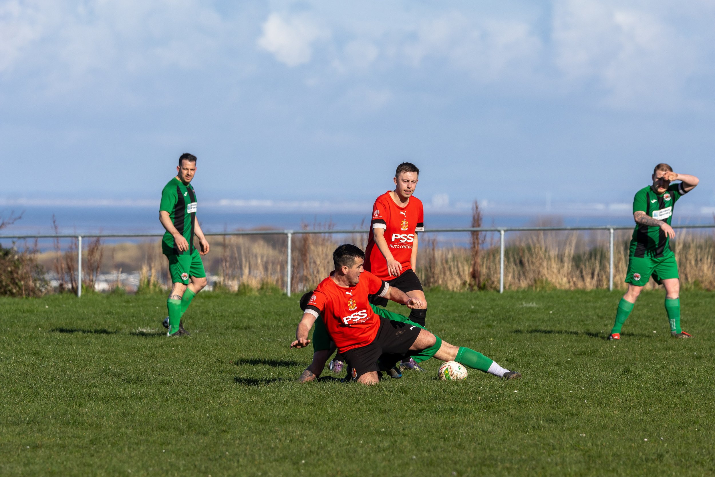 Four men playing soccer on a grassy field, two wearing red jerseys and two in green jerseys, with a landscape of water and distant land in the background.
