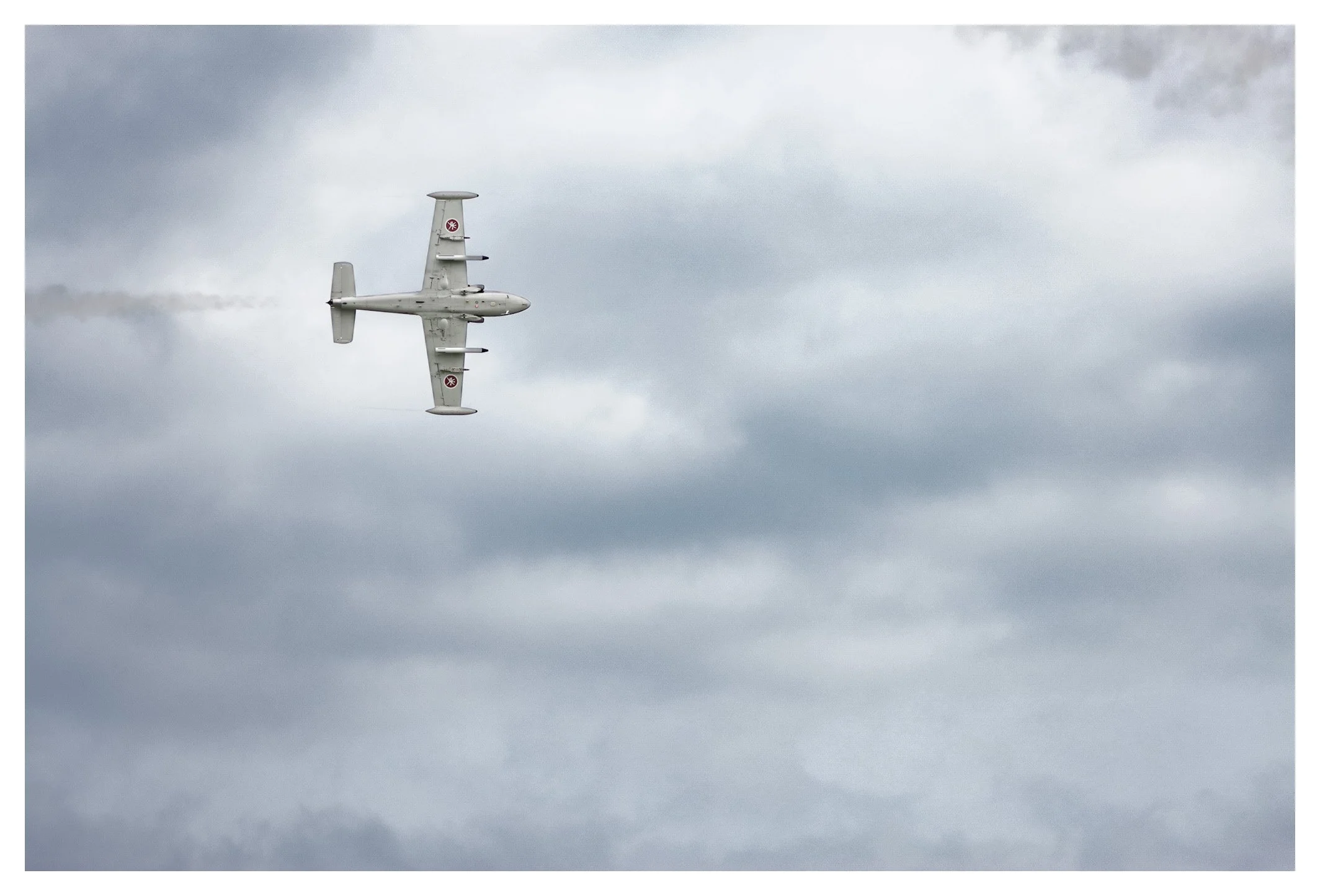 A military aircraft flying in cloudy sky, leaving a smoke trail behind.