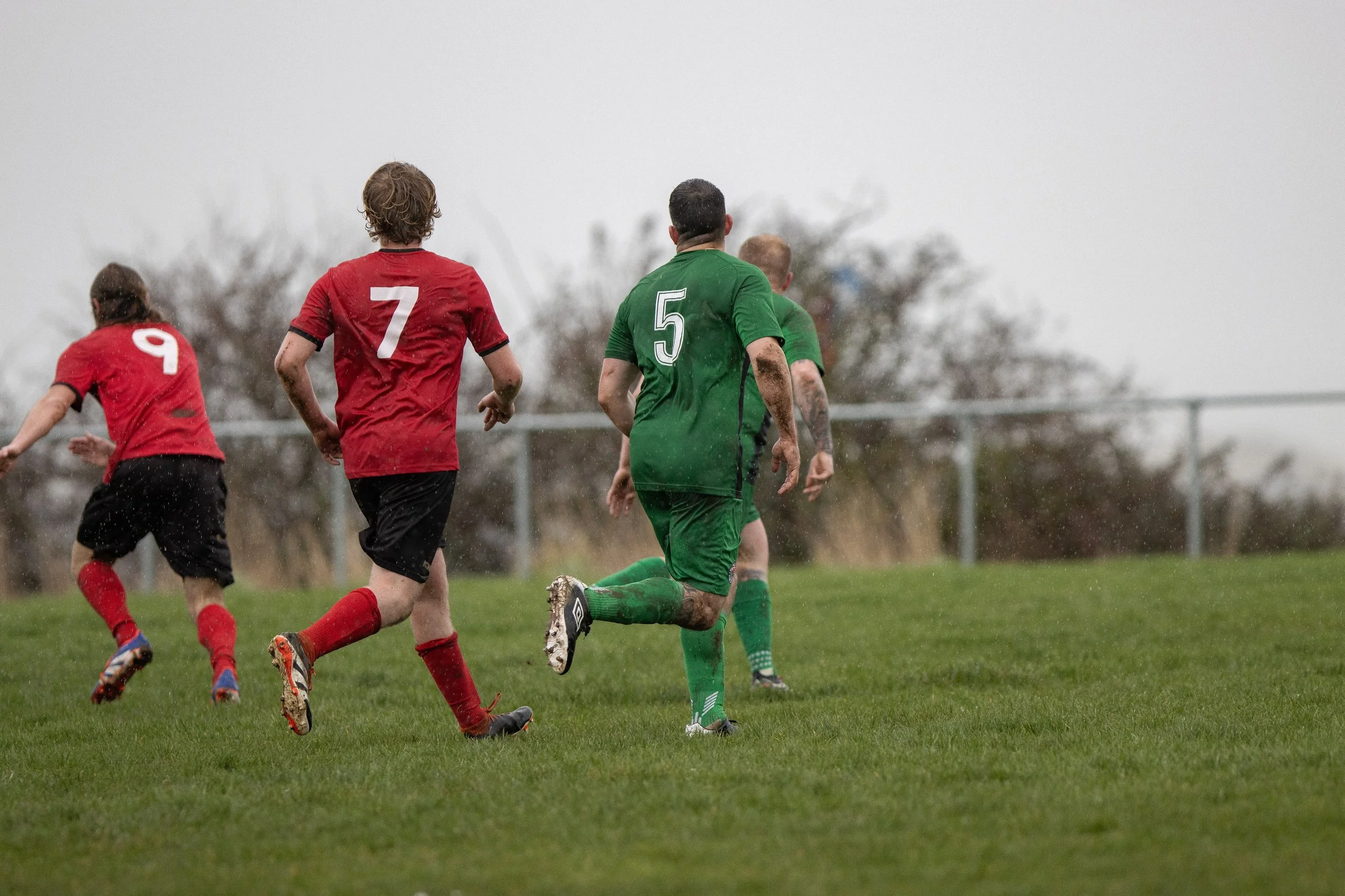 Soccer players running on a wet grassy field during a game, wearing red and green uniforms, with rain falling and trees and a fence in the background.